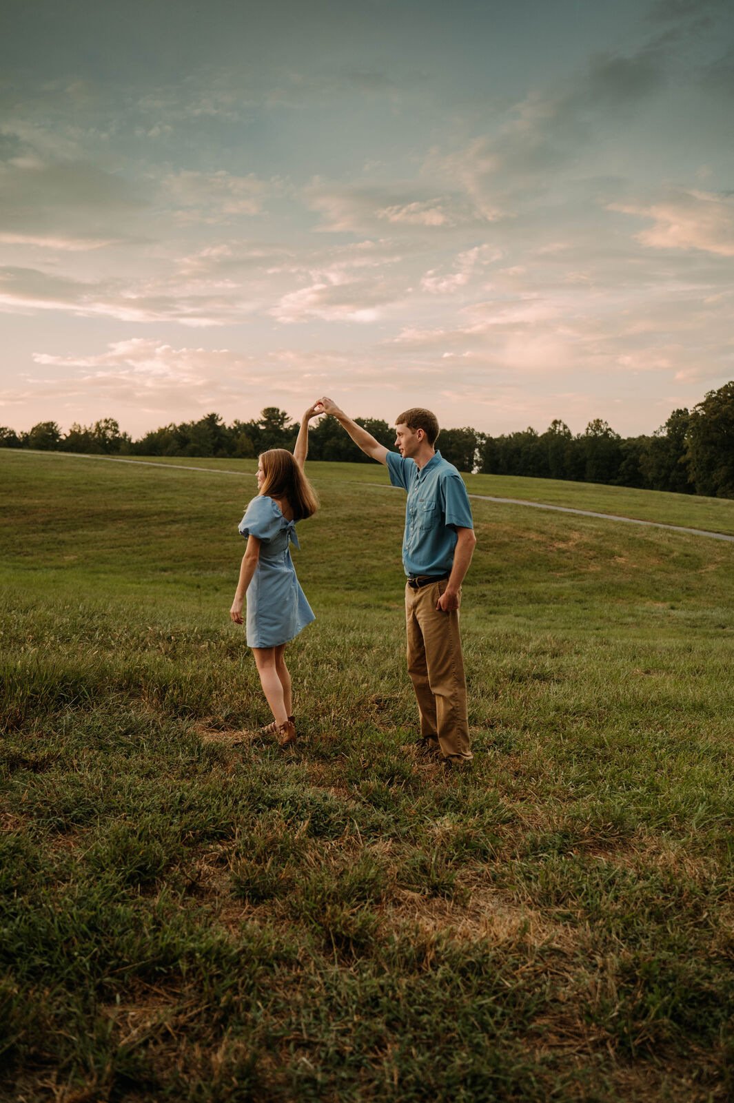 A young man and woman dance together outdoors in a grassy field during sunset, with the man twirling the woman, both wearing casual blue clothing.