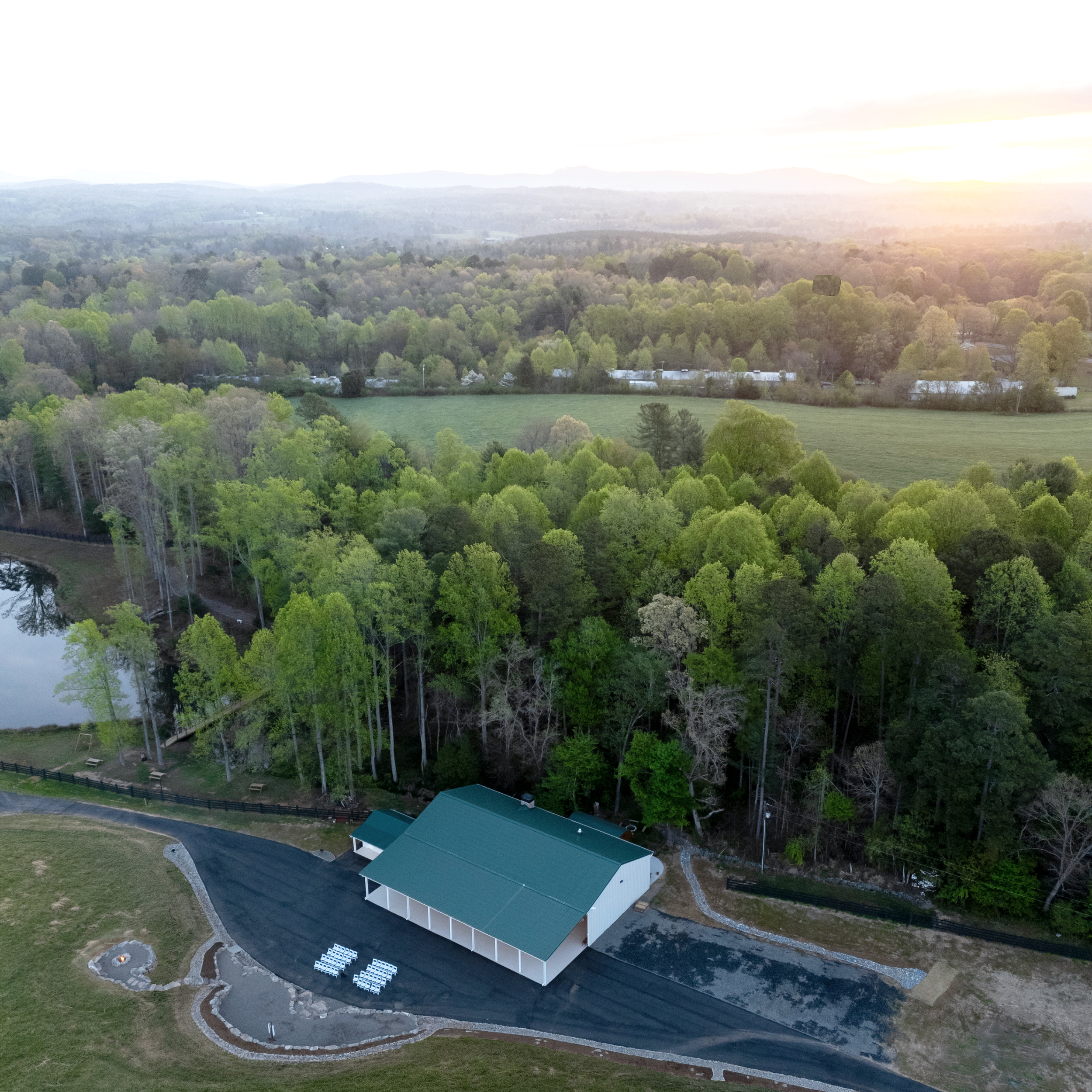 Aerial view of a landscape with green trees, a body of water, a small building, and hills in the distance during sunset.