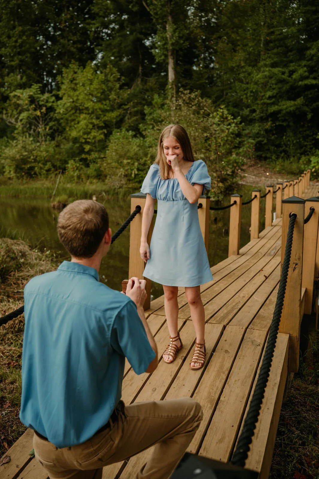A man kneeling on a wooden bridge proposing to a woman standing on the bridge in a forested area.