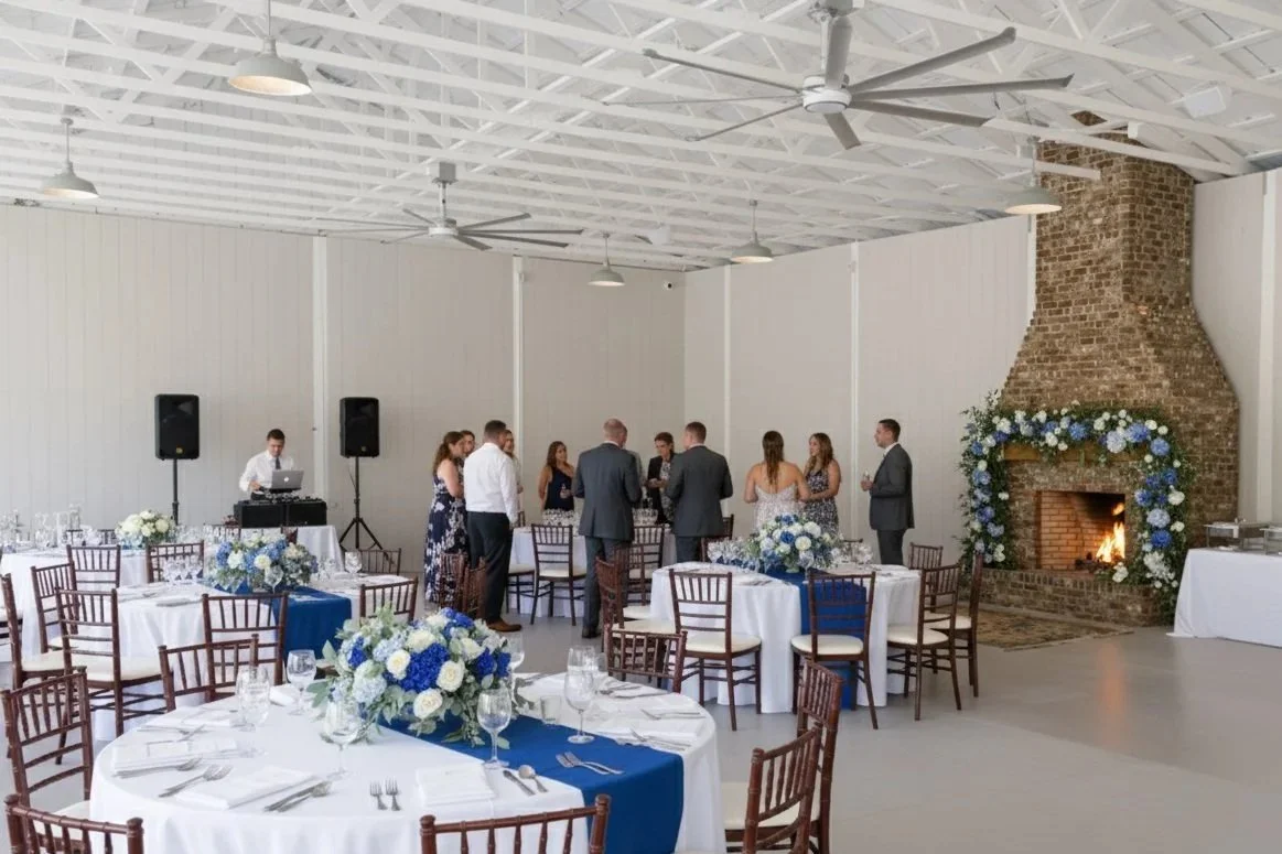 Wedding reception with round tables decorated with white tablecloths and blue runners, floral centerpieces with blue and white flowers, a brick fireplace with blue and white floral garland, people in semi-formal attire, and a DJ setup with speakers in a spacious hall with white walls and ceiling fans.