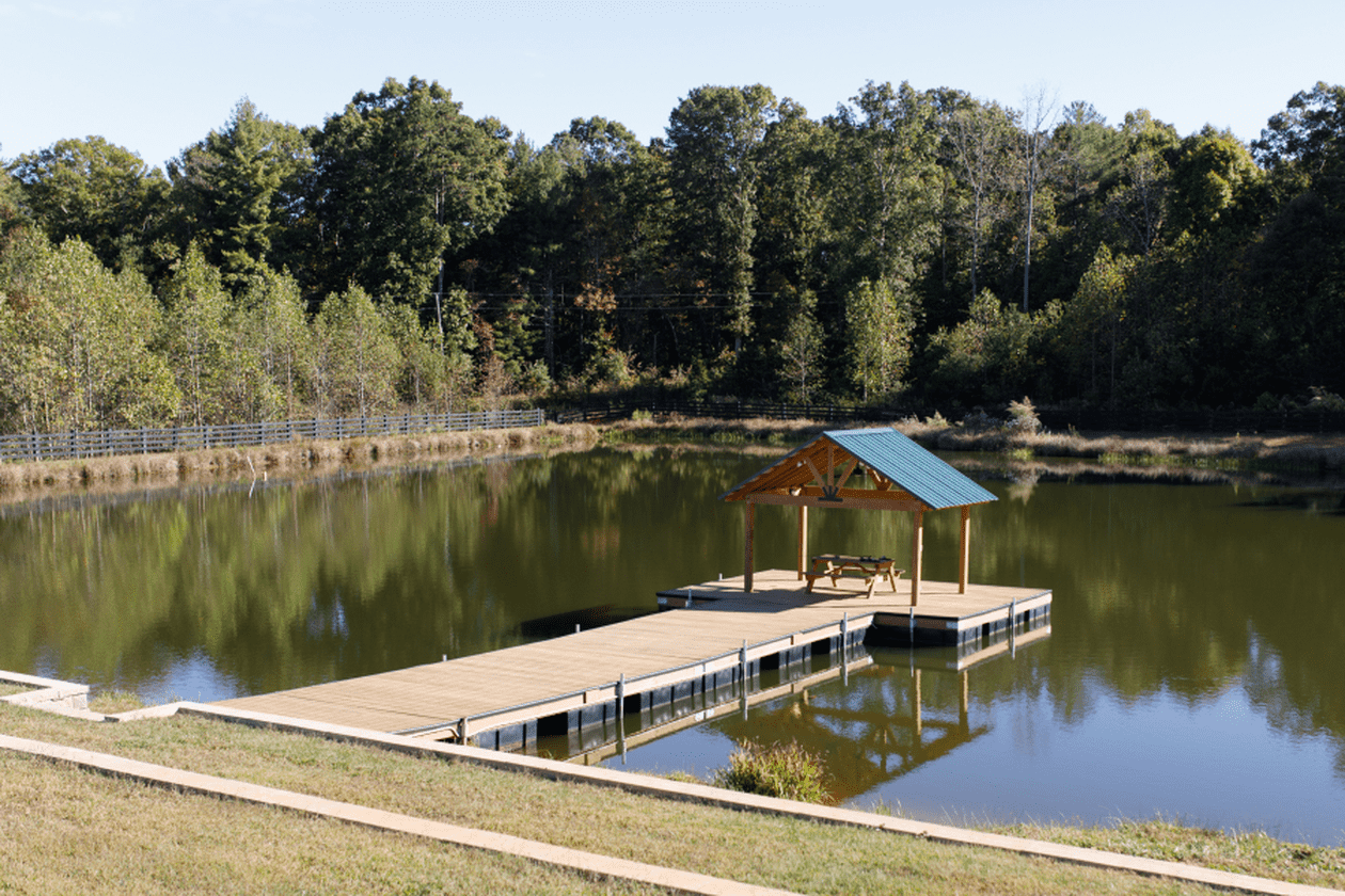 A wooden dock with a covered seating area extending over a calm lake, surrounded by trees and a grassy shoreline.