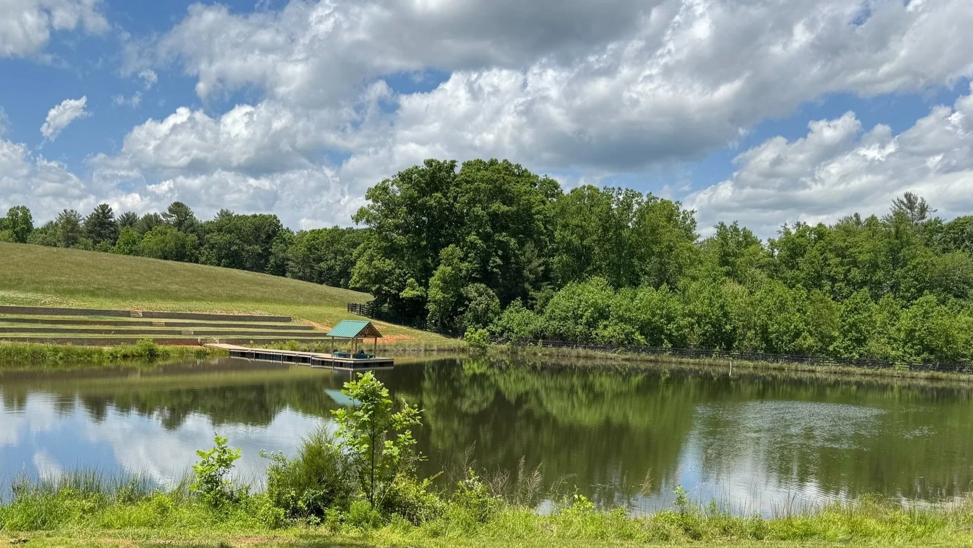 A serene pond with a small dock featuring a shaded shelter, surrounded by green trees and rolling hills under a partly cloudy sky.