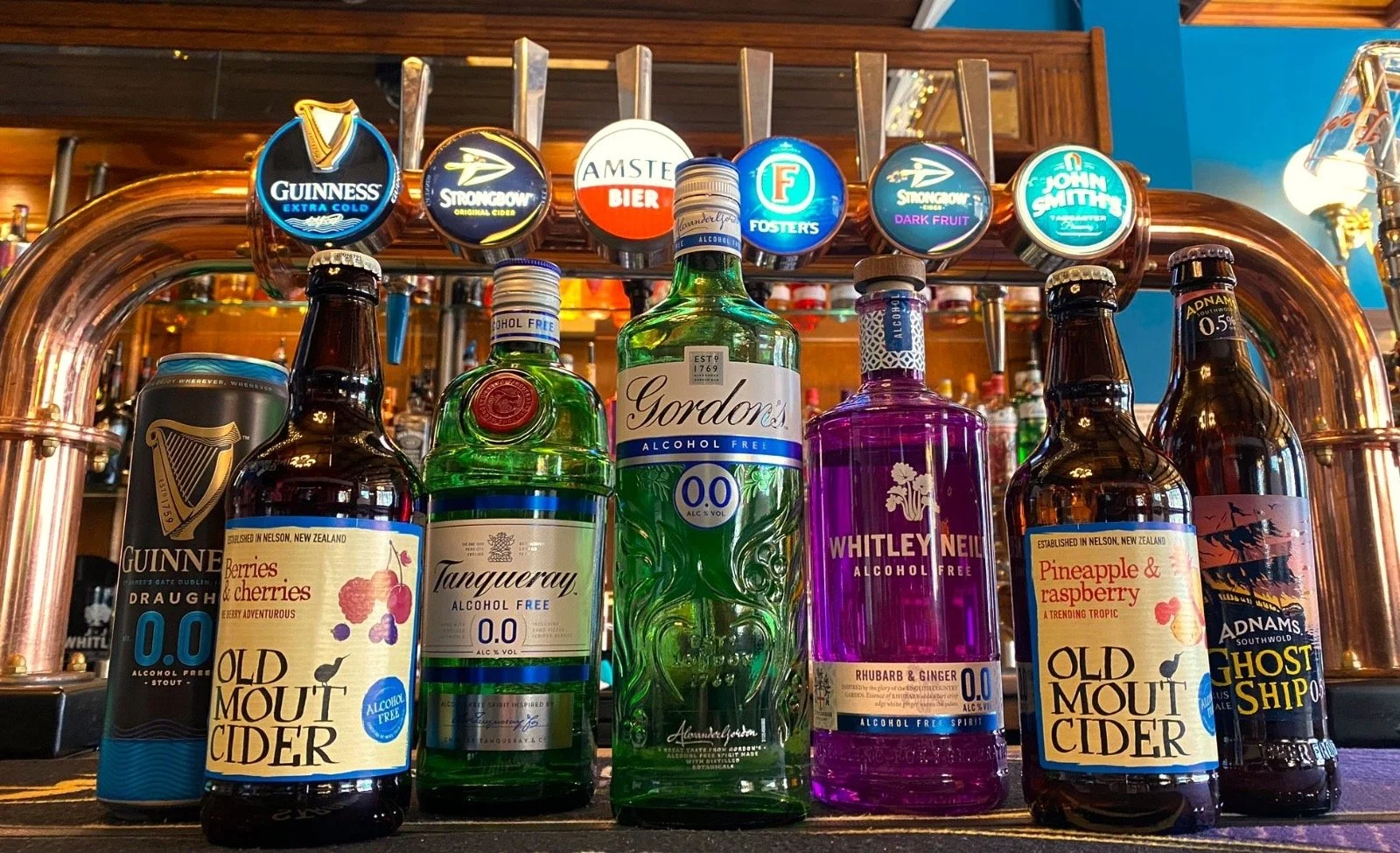 A selection of alcohol-free beverages, including cans and bottles of cider, mocktails, and flavored spirits, displayed on a bar with beer tap handles featuring various beer brands and flavors.