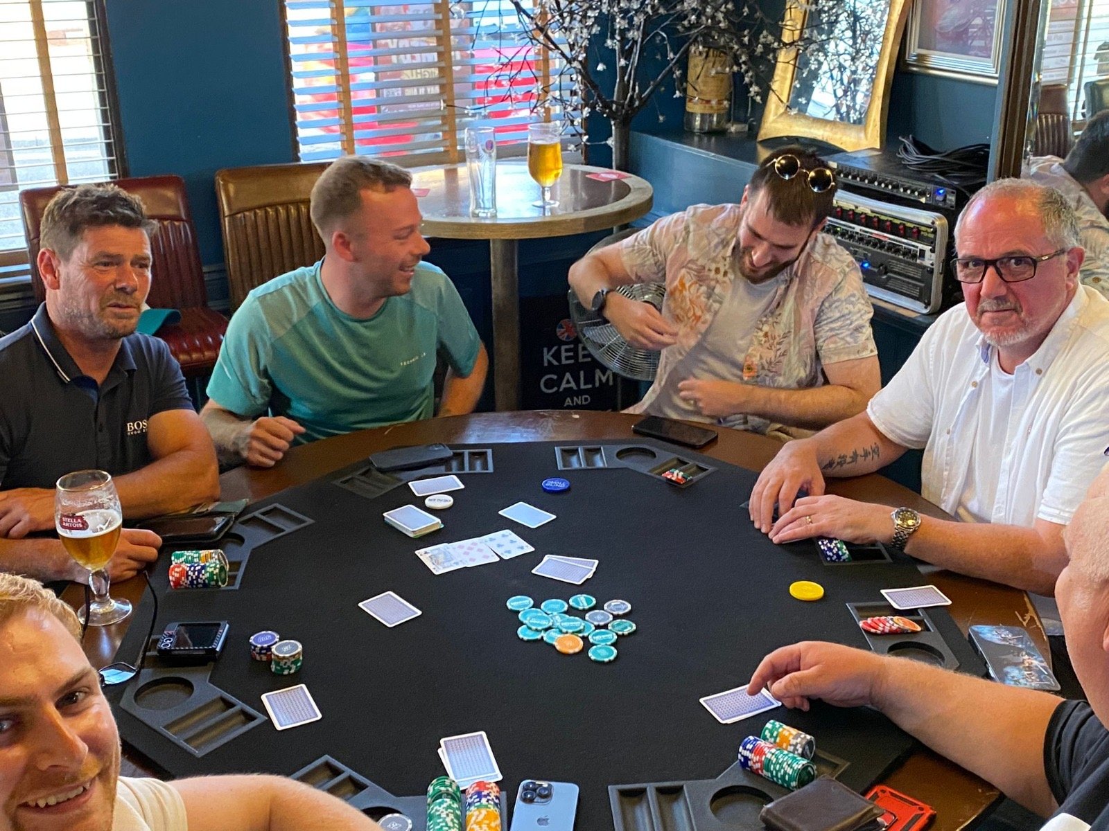 A group of six men playing poker at a round table with chips and cards, in a restaurant or pub setting.