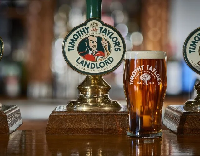A draft beer tap labeled Timothy Taylor's Landlord above a pint glass filled with amber beer, on a wooden bar counter.