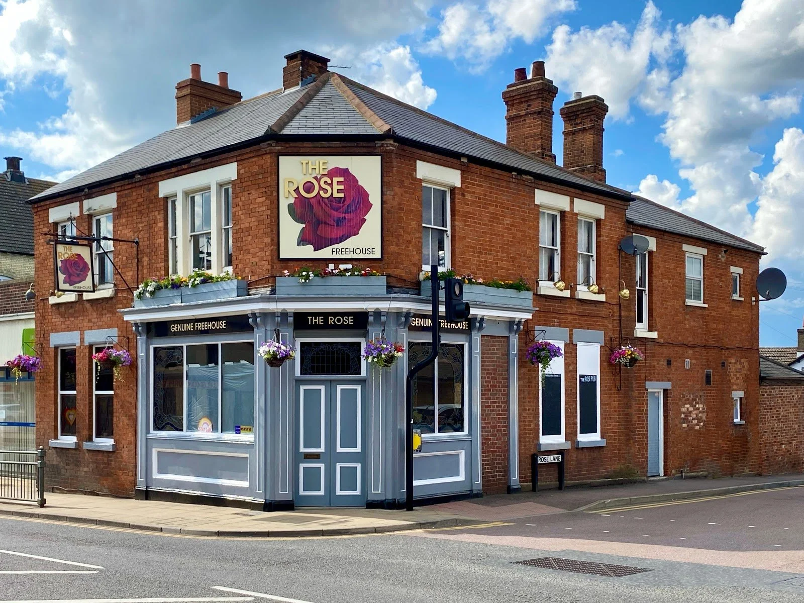 A red brick building named The Rose pub with hanging flower baskets and a large sign featuring a purple rose, situated on a street corner with a blue sky and clouds above.