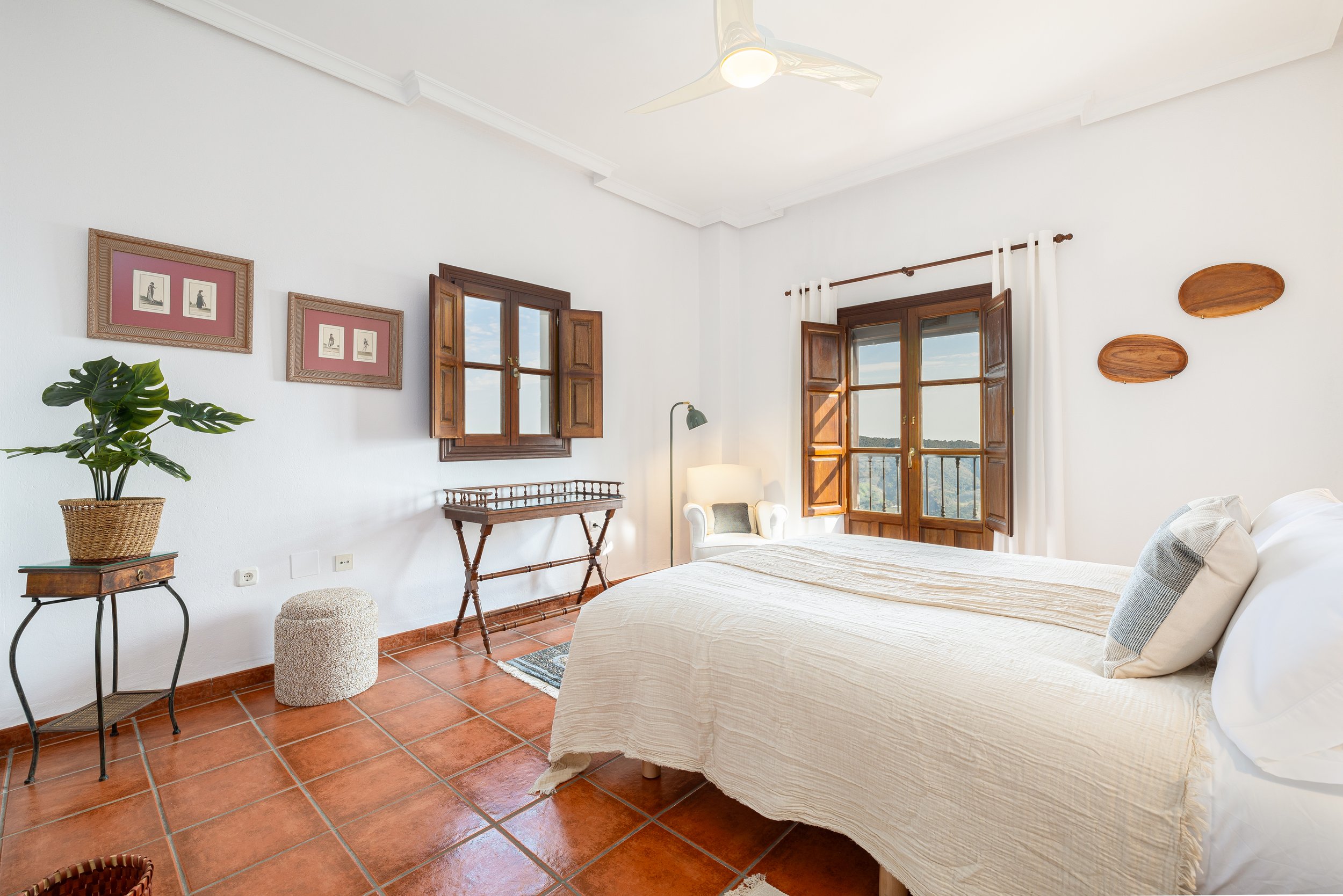 Bright bedroom with white walls, terracotta tile floor, and wooden-framed windows showing a scenic view. Contains a bed with beige bedding, a white armchair, a small side table with a potted plant, framed artwork on the wall, and rustic wooden wall decor.