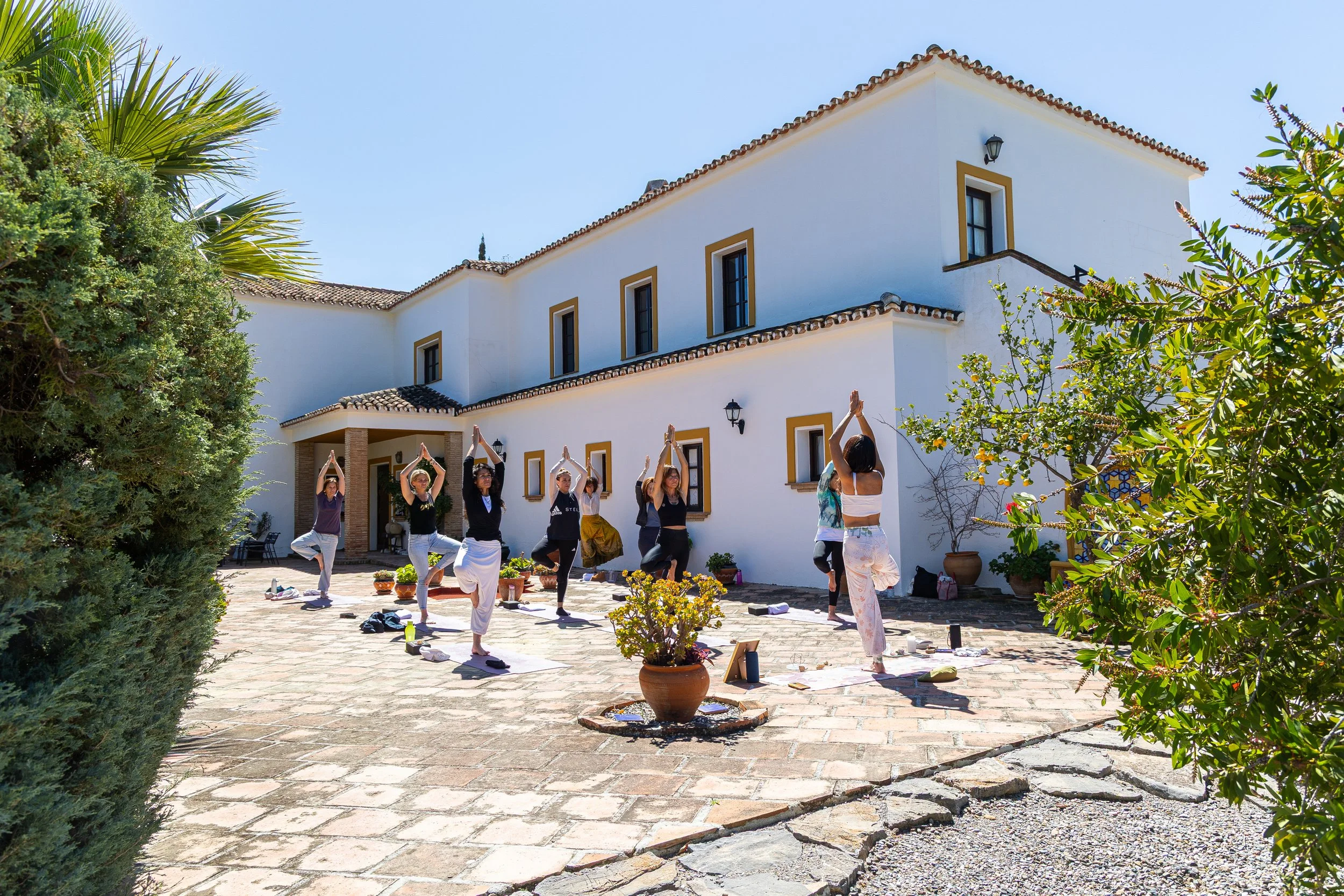 Group of people practicing yoga outdoors on a patio in front of a white Mediterranean-style house under a clear blue sky.