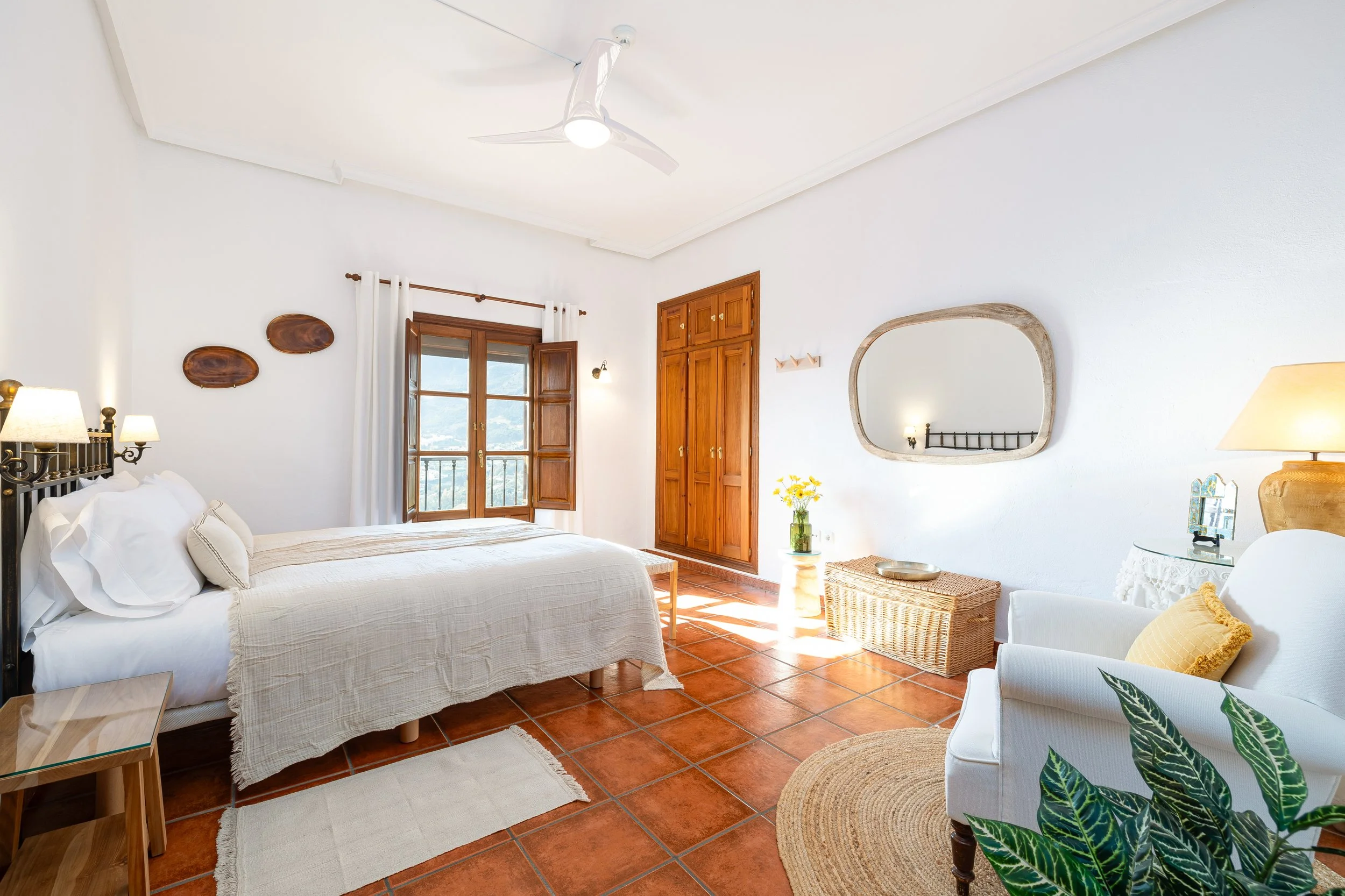 A bedroom with a white bed, wooden furniture, terracotta floor tiles, and natural light from open windows with white curtains, decorated with cozy furniture and plants.