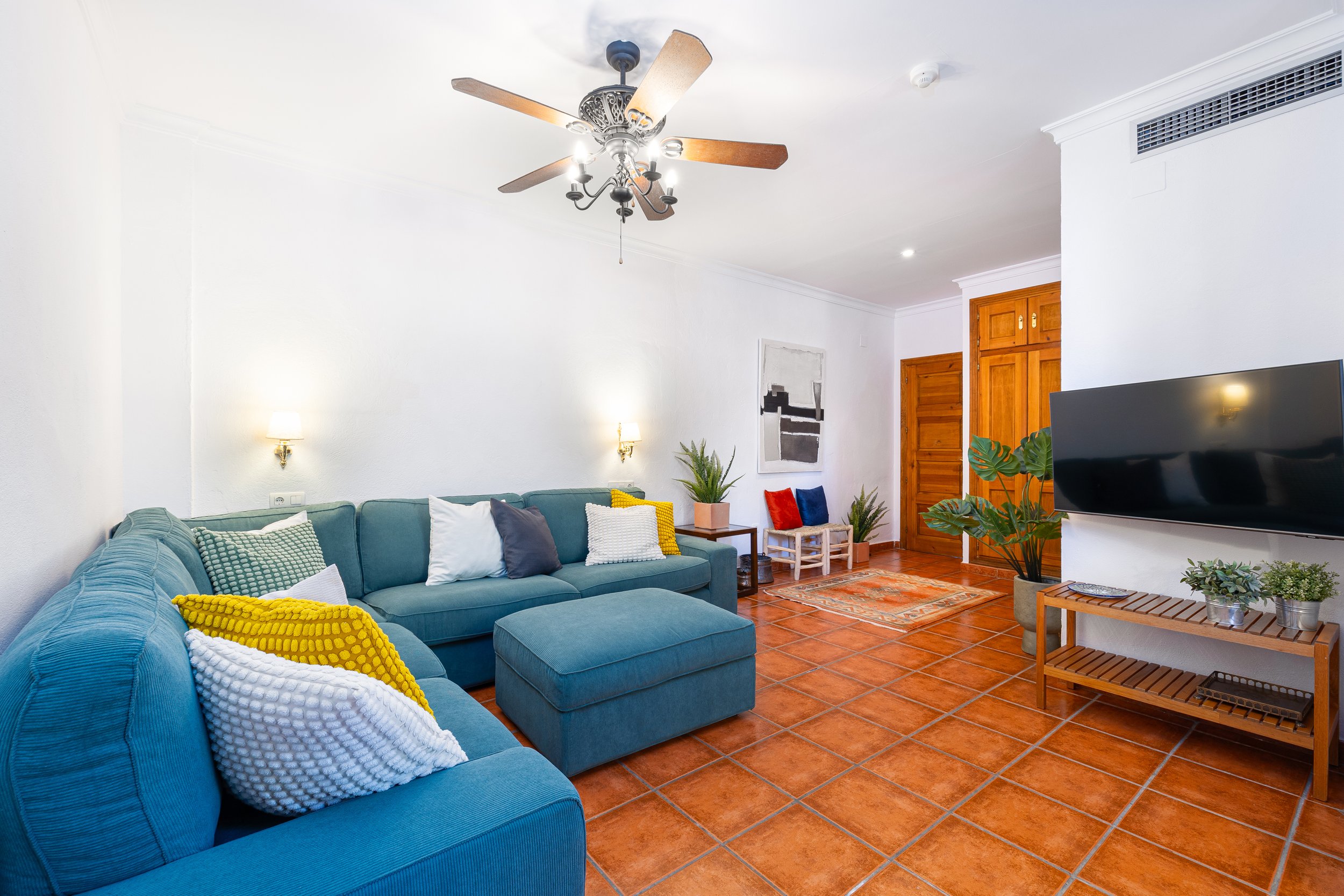 Living room with white walls, wooden doors, a blue sectional sofa with colorful pillows, a ceiling fan, wall-mounted TV, and various potted plants.