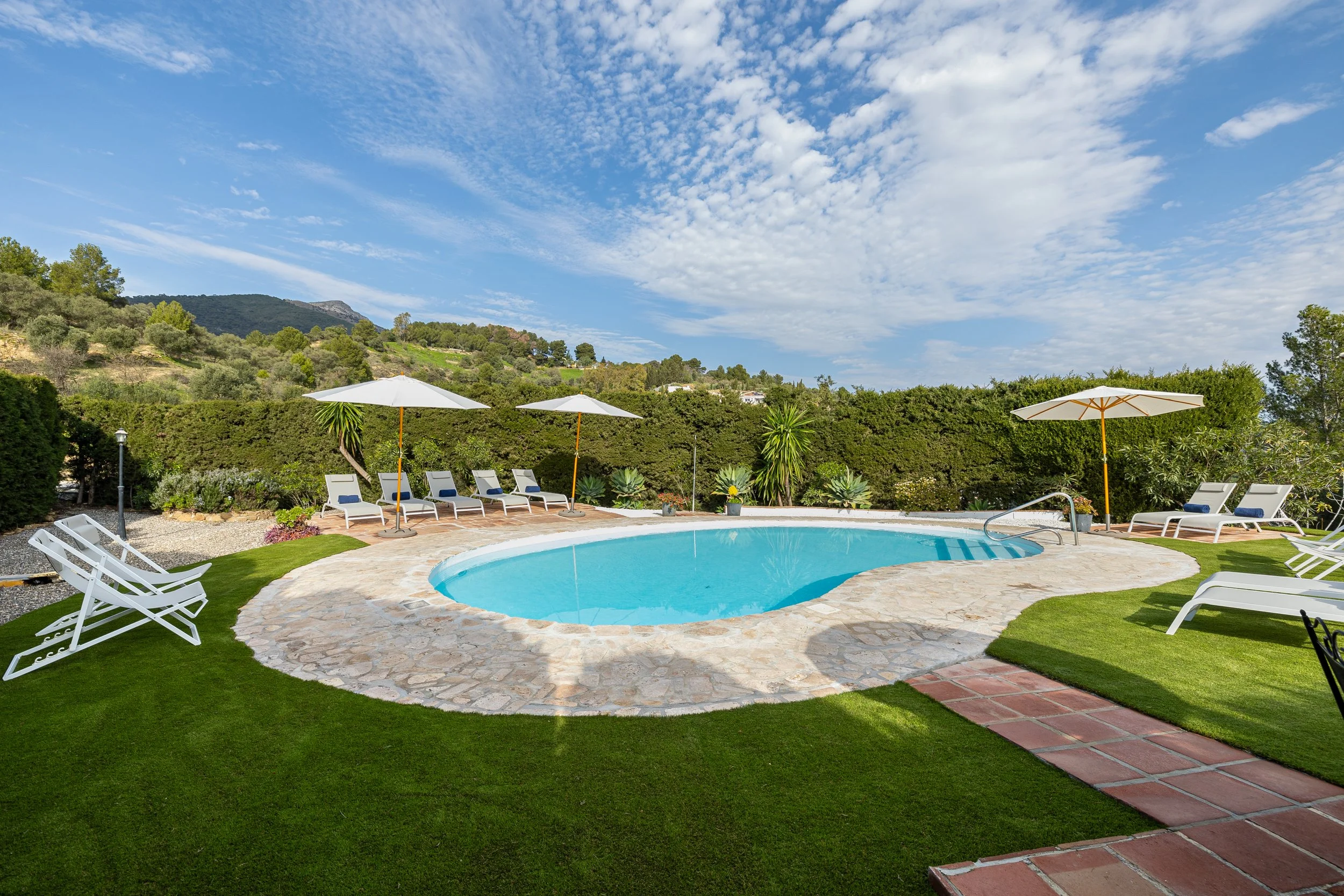 Outdoor swimming pool with lounge chairs, umbrellas, and lush greenery under a partly cloudy sky.