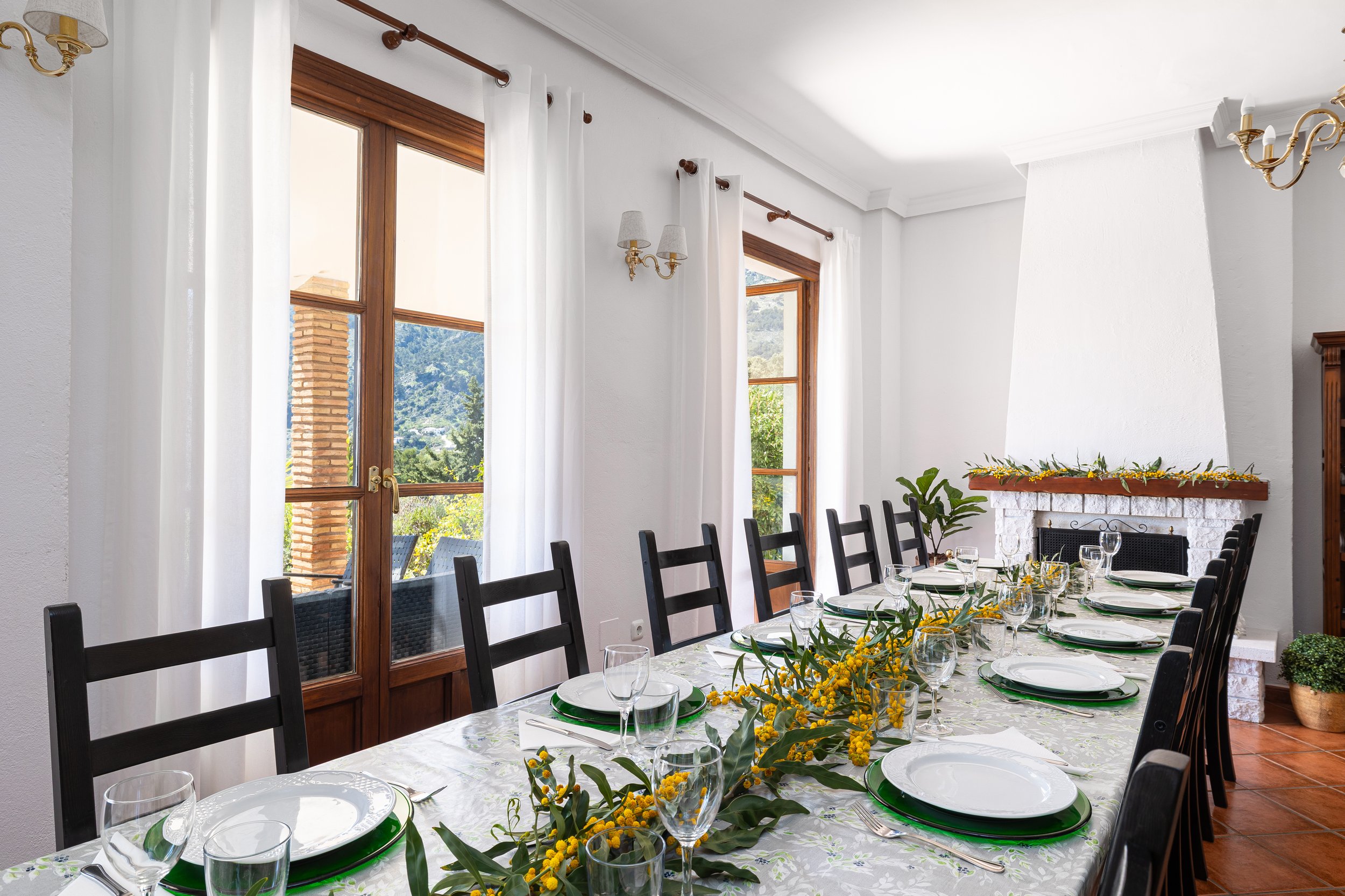 A dining room with a long table set for a meal, decorated with green and yellow floral arrangements, with black chairs, white plates, and wine glasses. Large windows with white curtains let in natural light, and a white fireplace with a wooden mantel and a plant inside. The room has tiled floors and white walls.