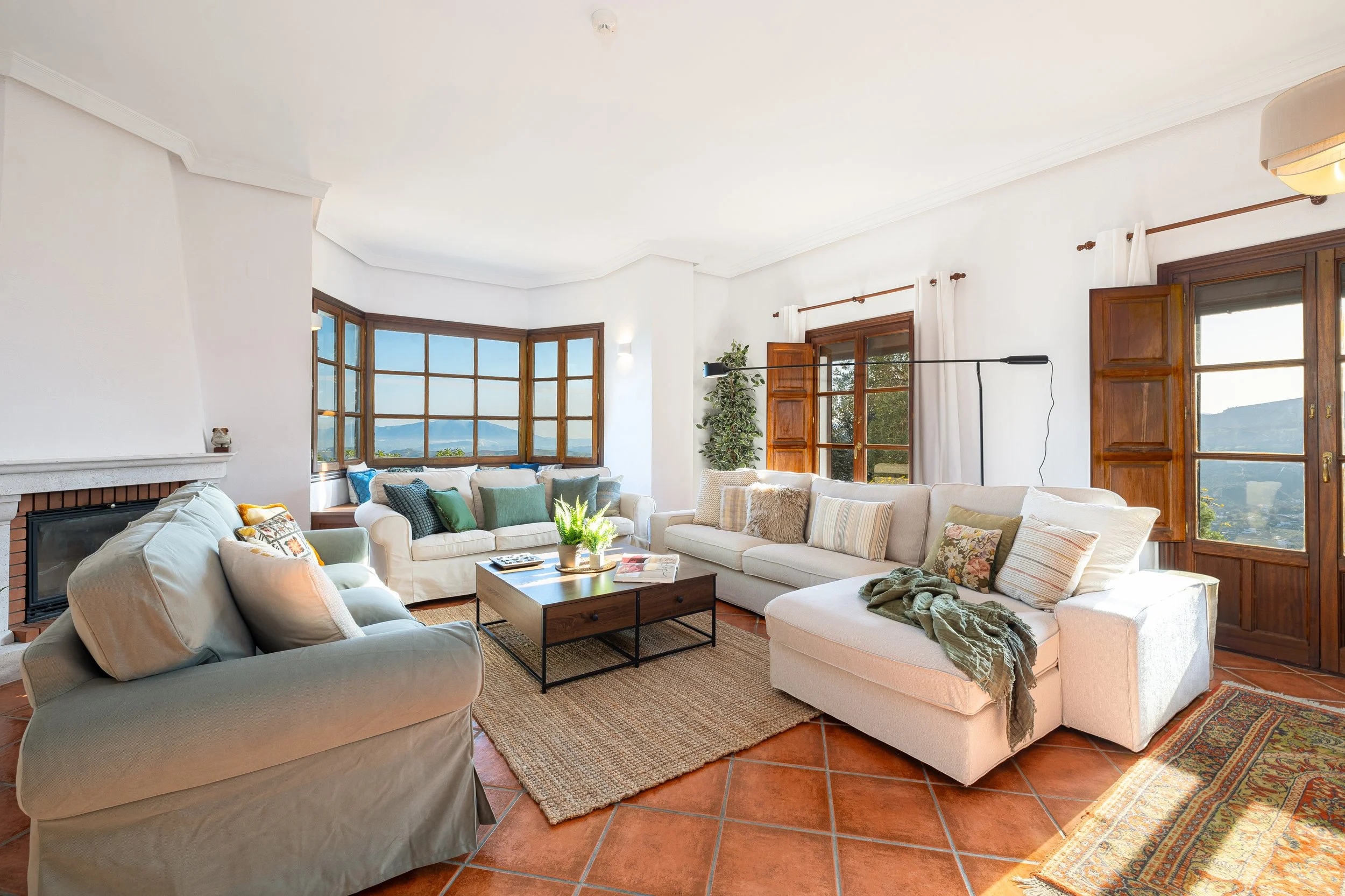 Bright living room with large windows, cream-colored sectional sofa, wood coffee table, and terracotta tile floor.
