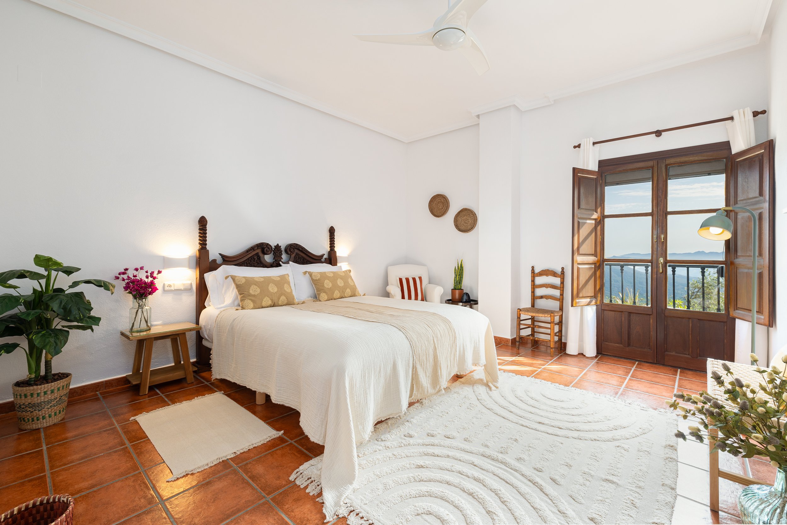 Bright bedroom with white walls, terracotta tile flooring, a carved wooden bed with white linens and beige pillows, a potted plant on the side table, a white armchair with striped cushion, wooden chairs, a large window with wooden shutters and a scenic mountain view, and a cream rug.