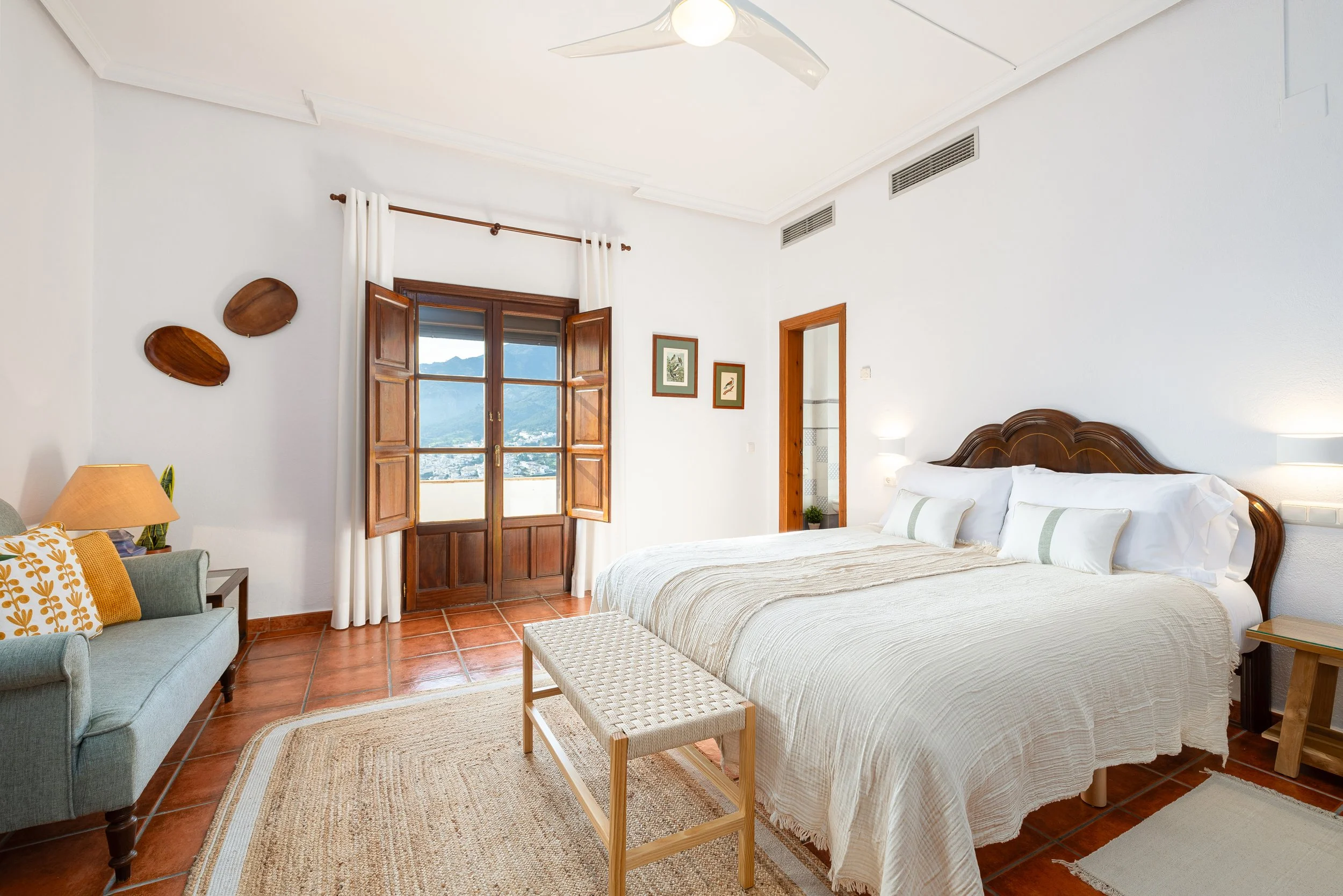 Sunlit bedroom with large bed, wooden headboard, white bedding, pillows, a small bench at foot of bed, terracotta tile floor, green sofa with patterned pillow, wooden side tables, window with wooden shutters, mountain view, framed pictures on wall, white walls, ceiling fan with light.