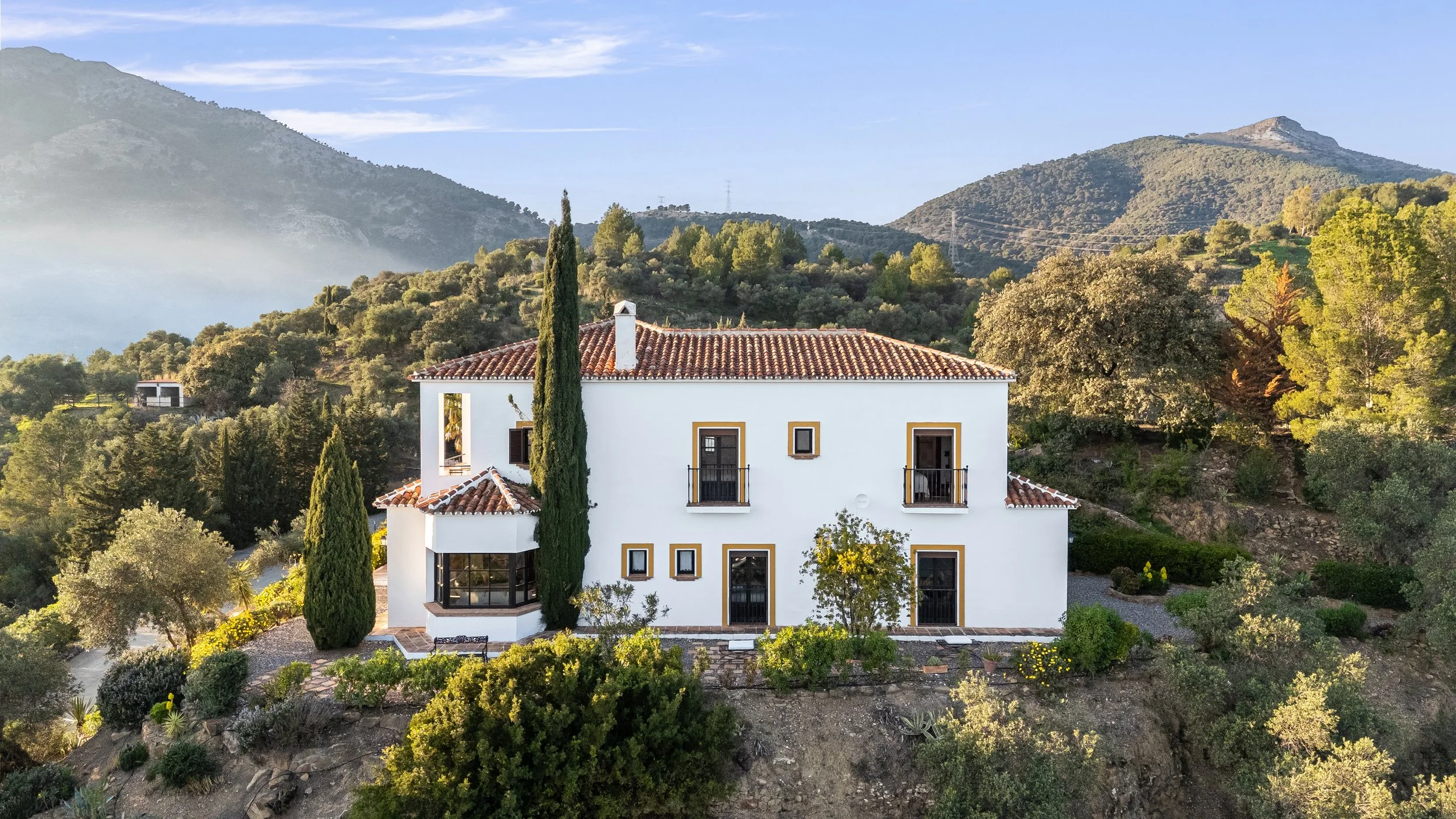 A white house with a red-tile roof surrounded by greenery and trees, set against a backdrop of hills and mountains under a partly cloudy sky.