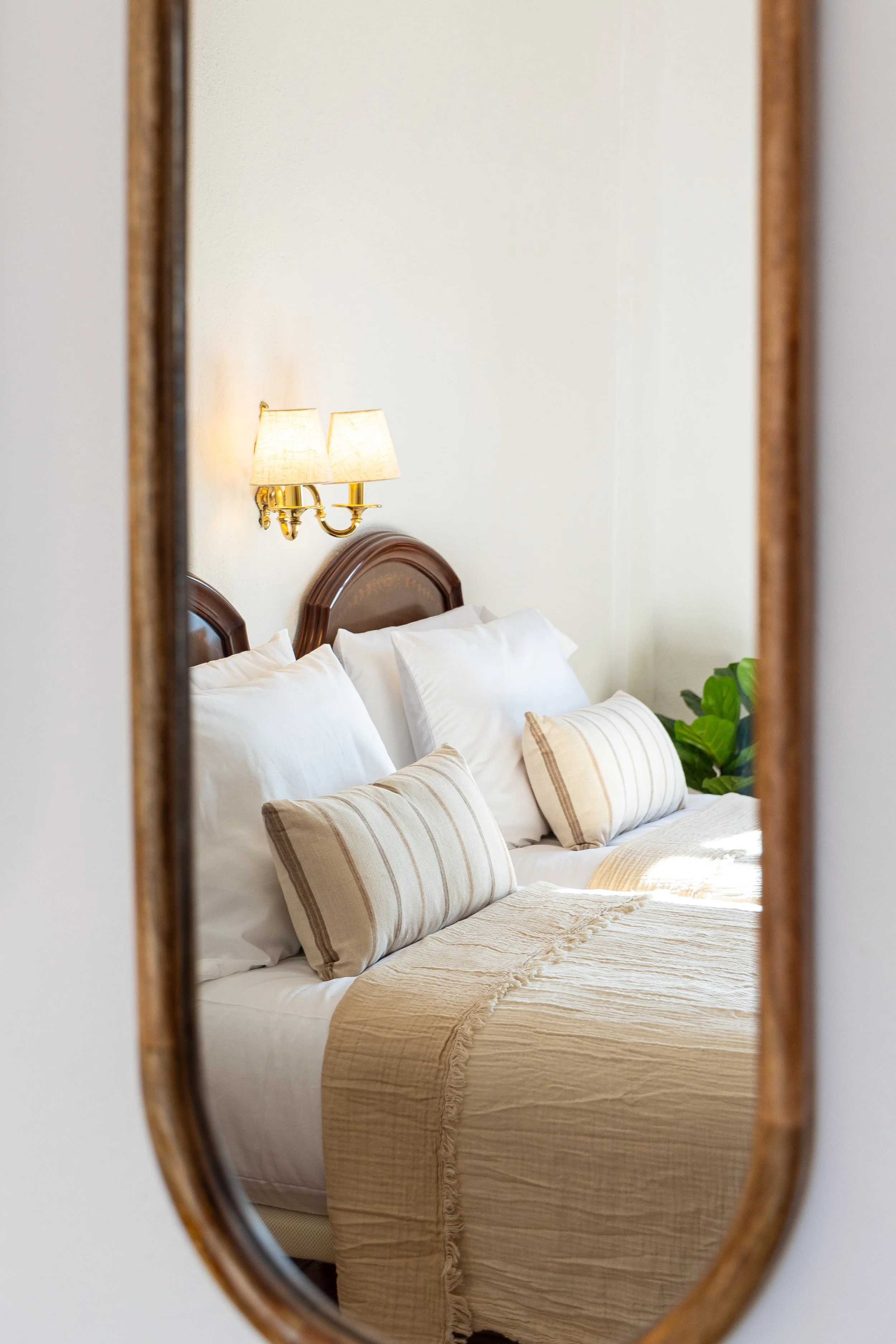 Reflection in a mirror showing a neatly made bed with white pillows, beige striped cushions, beige bedspread, wooden headboard, wall-mounted lamps, and green plant in a cozy bedroom.