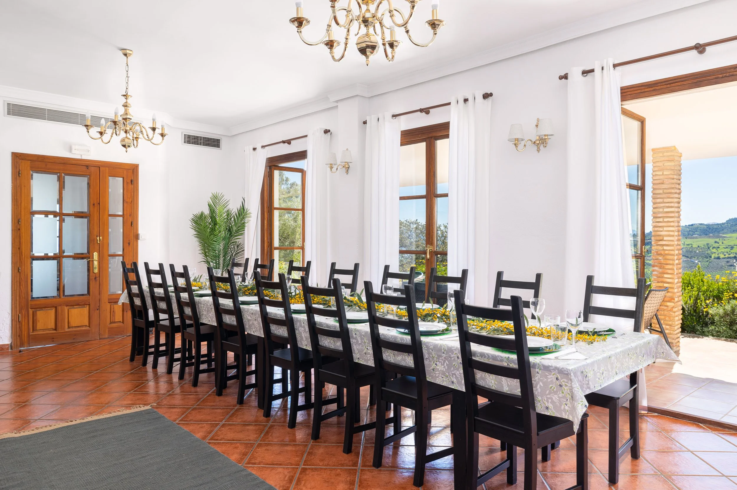 A spacious dining room with a long table covered with a floral tablecloth, set for a meal with plates, glasses, and a centerpiece. The room has large windows with white curtains, wooden frames, and a view of green hills outside. There are chandeliers and wall-mounted lights, and terracotta tile flooring.