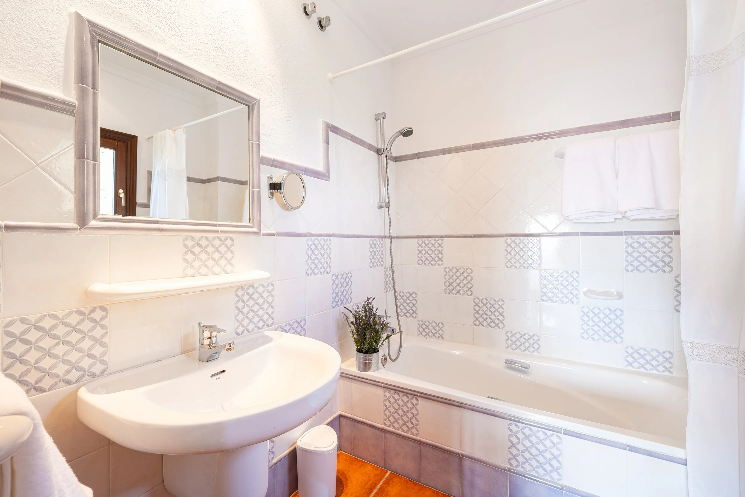 A bright bathroom with a white sink, mirror, bathtub with a lavender border, and a showerhead. Towel rack with folded towels, small potted plant on the edge of the bathtub, and a window reflected in the mirror.