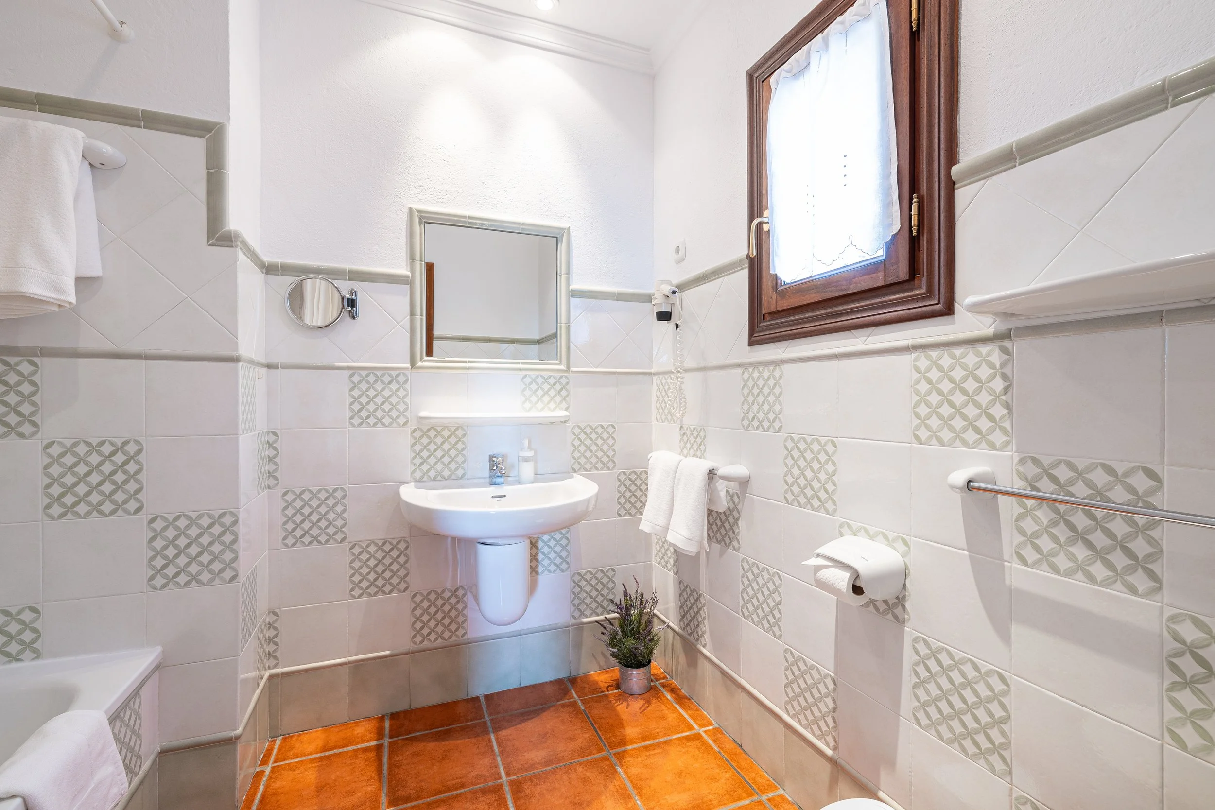 A small bathroom with white tiled walls, patterned tiles, a small sink with a mirror above it, and a window with a wooden frame and white curtain. There are towels, toilet paper, and a potted plant at the bottom.