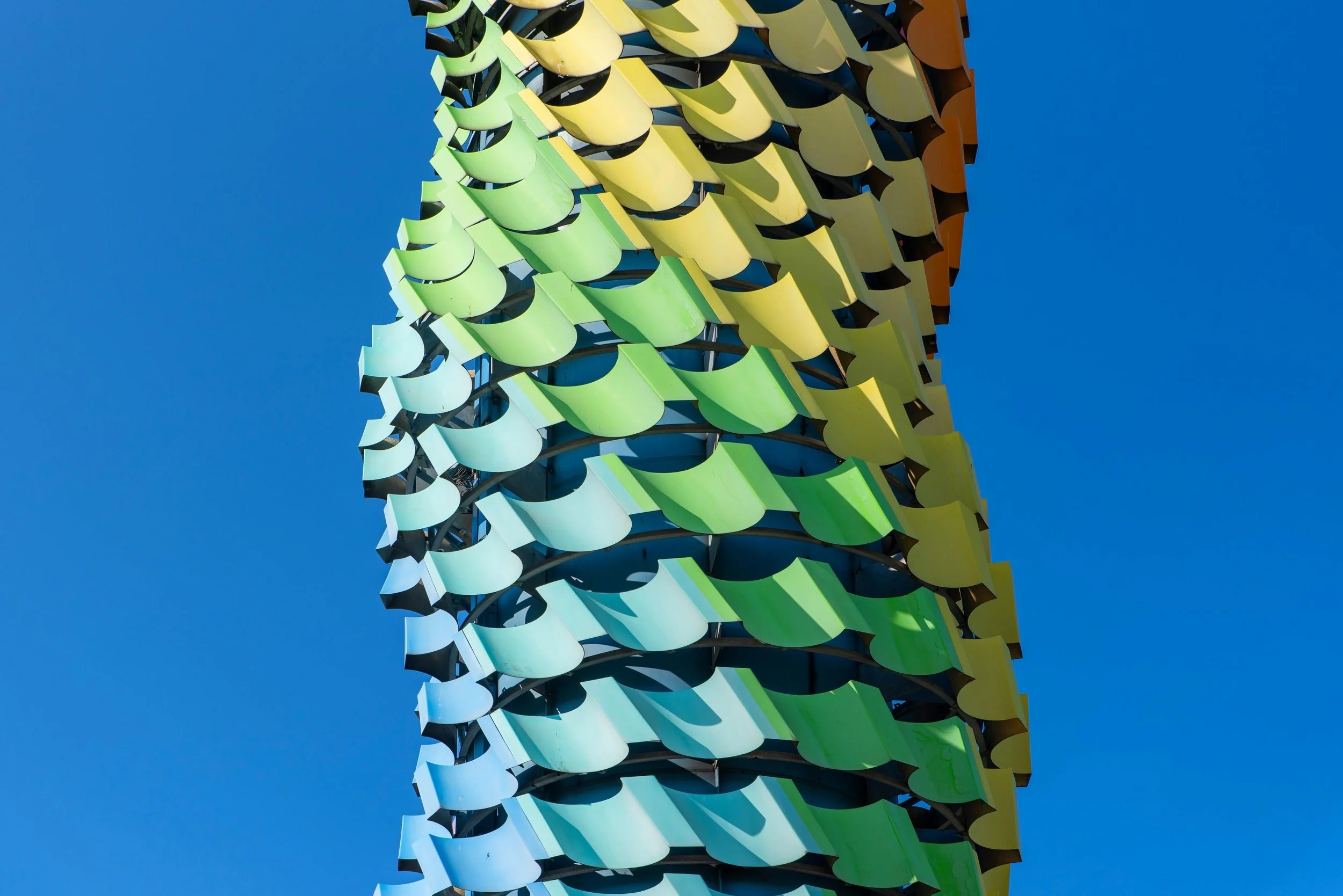 A colorful modern sculpture with a pattern of curved, overlapping panels in shades of green, yellow, and orange against a clear blue sky.