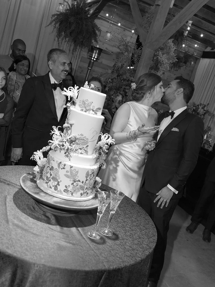 A wedding reception with a bride and groom sharing a kiss, a three-tier wedding cake decorated with floral patterns, and guests in the background smiling.