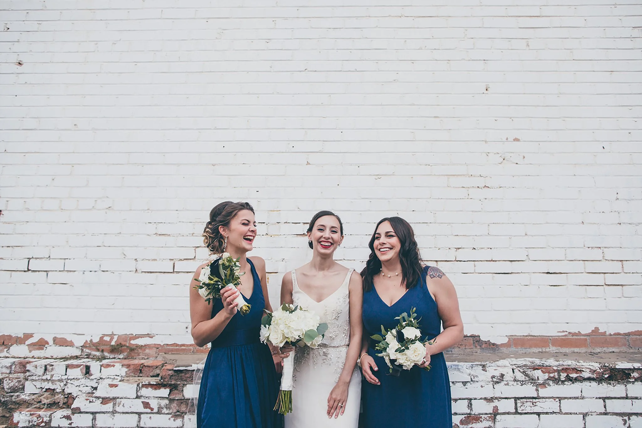 Carlisle Room wedding Bride and bridesmaids laughing against a whitewashed brick wall