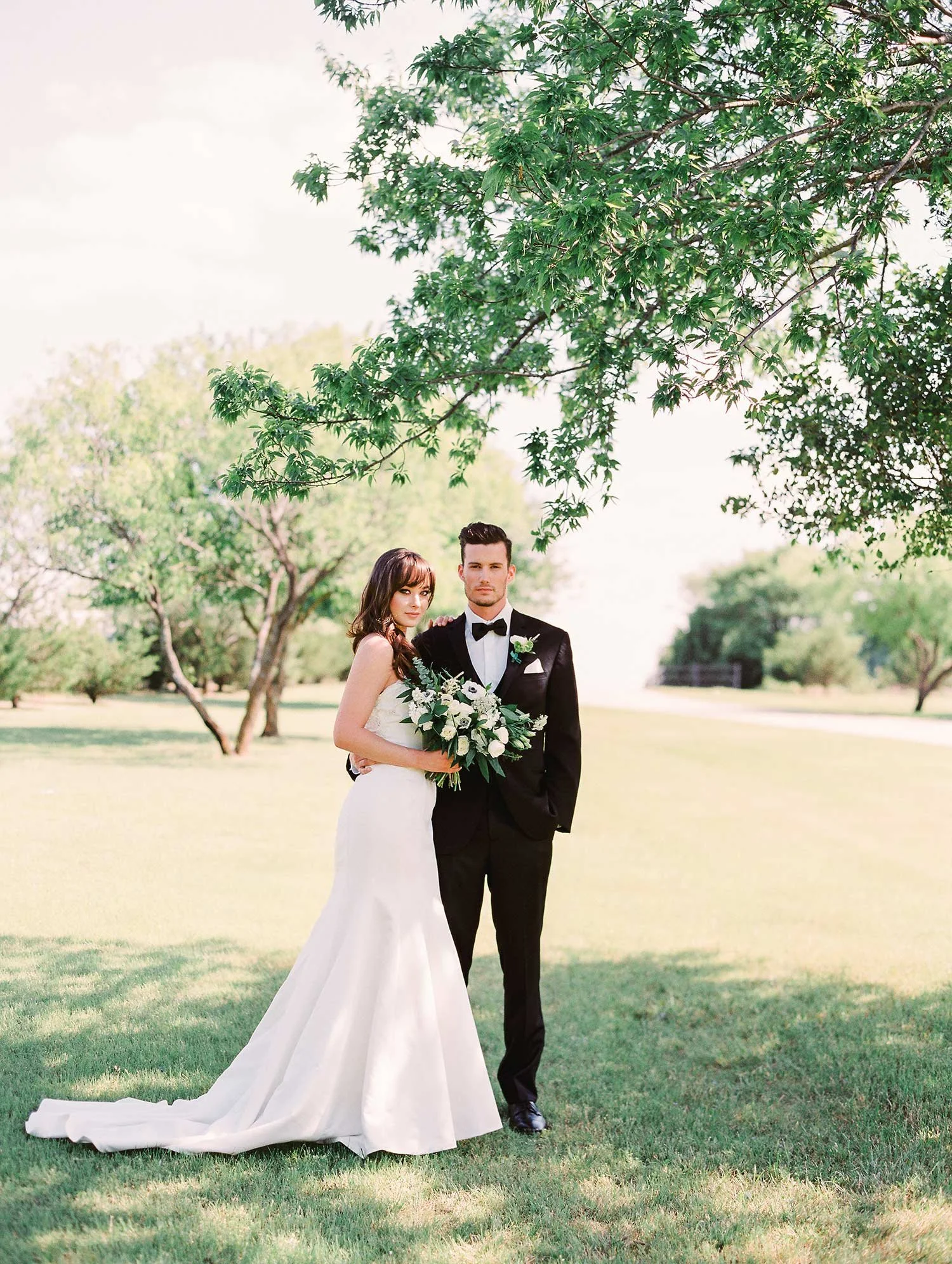 Bride in long satin dress standing outside with groom in black tuxedo