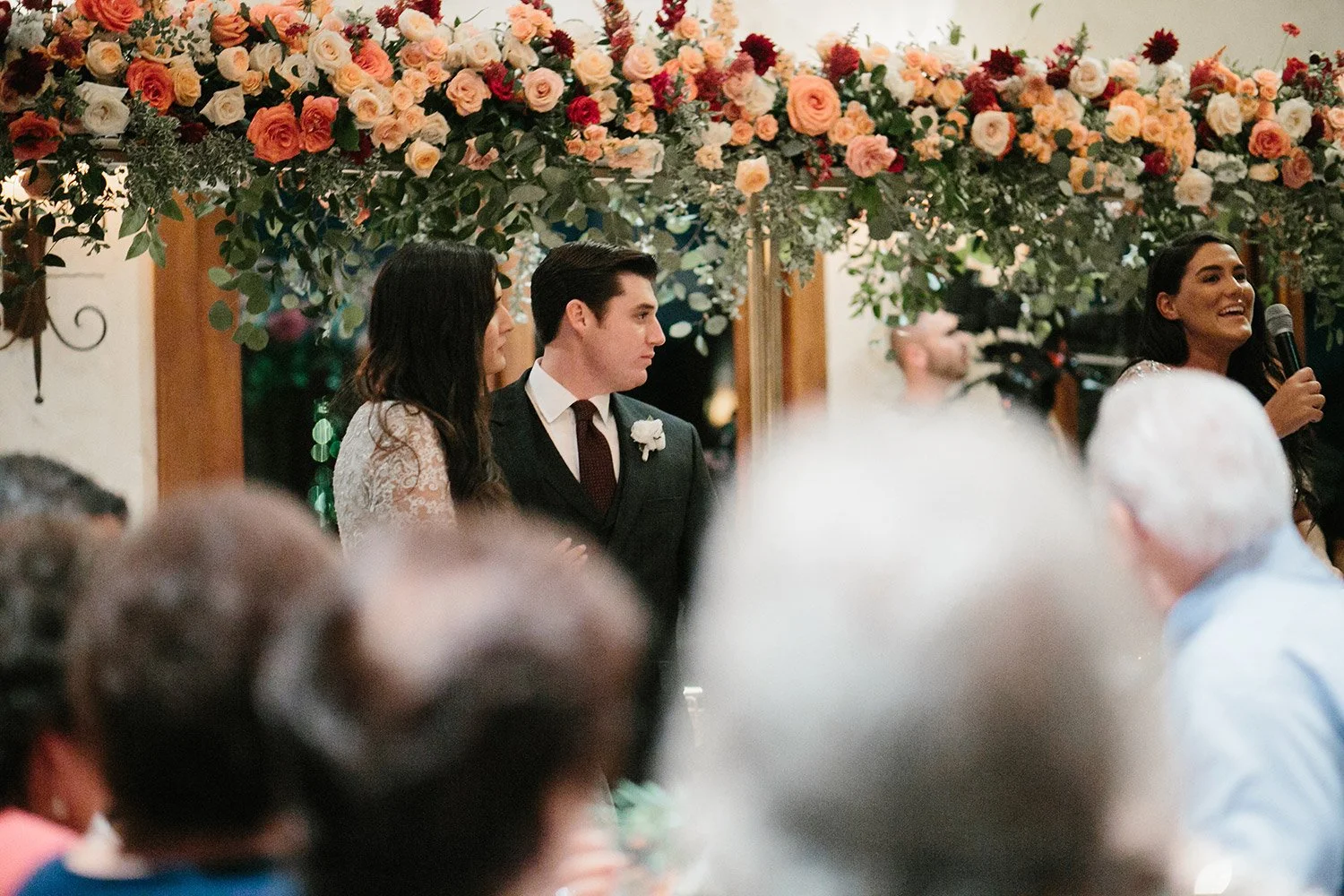 Bride and groom listening to speeches infront of tall flower garland