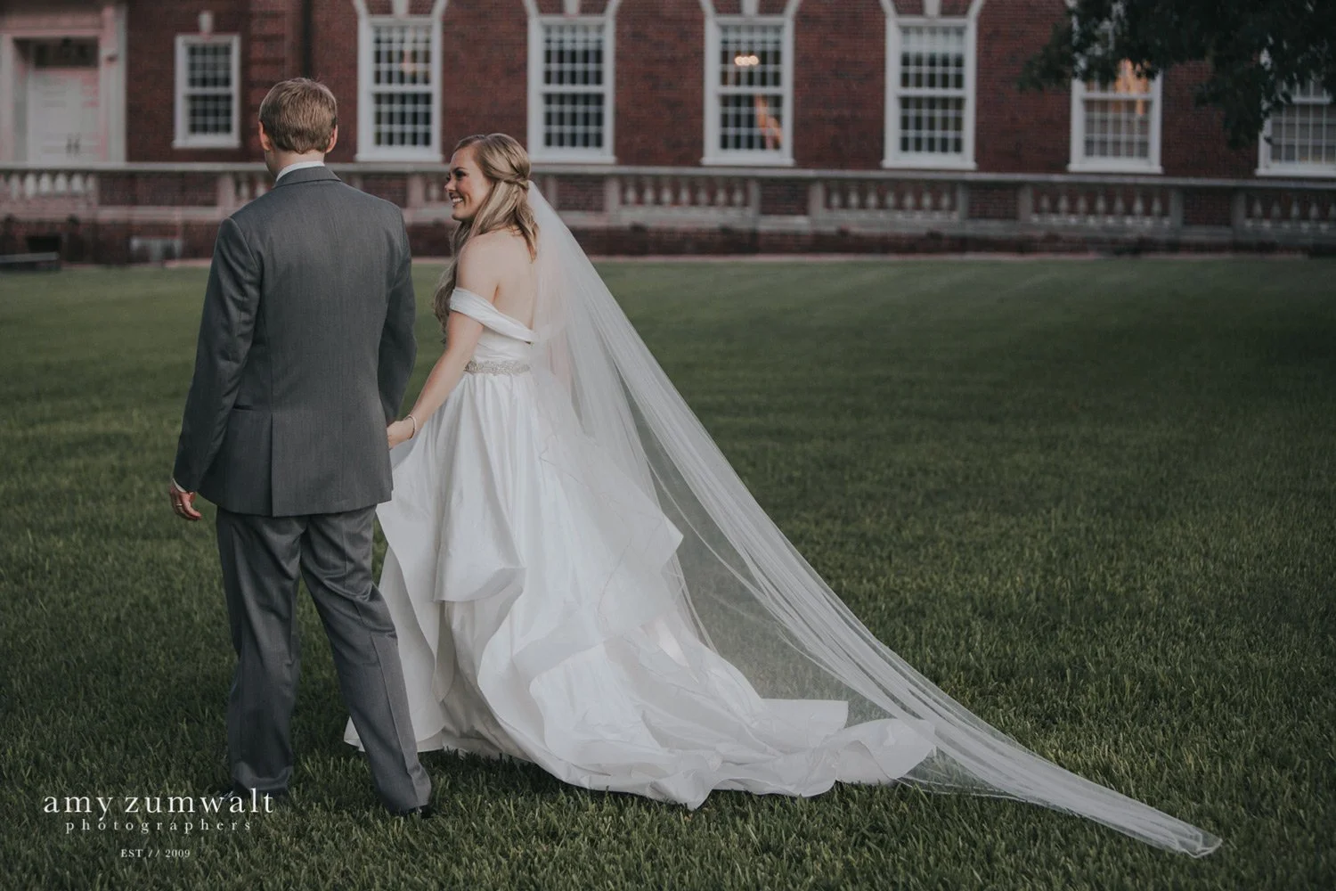 Bride and groom walking away in green field with cathedral veil