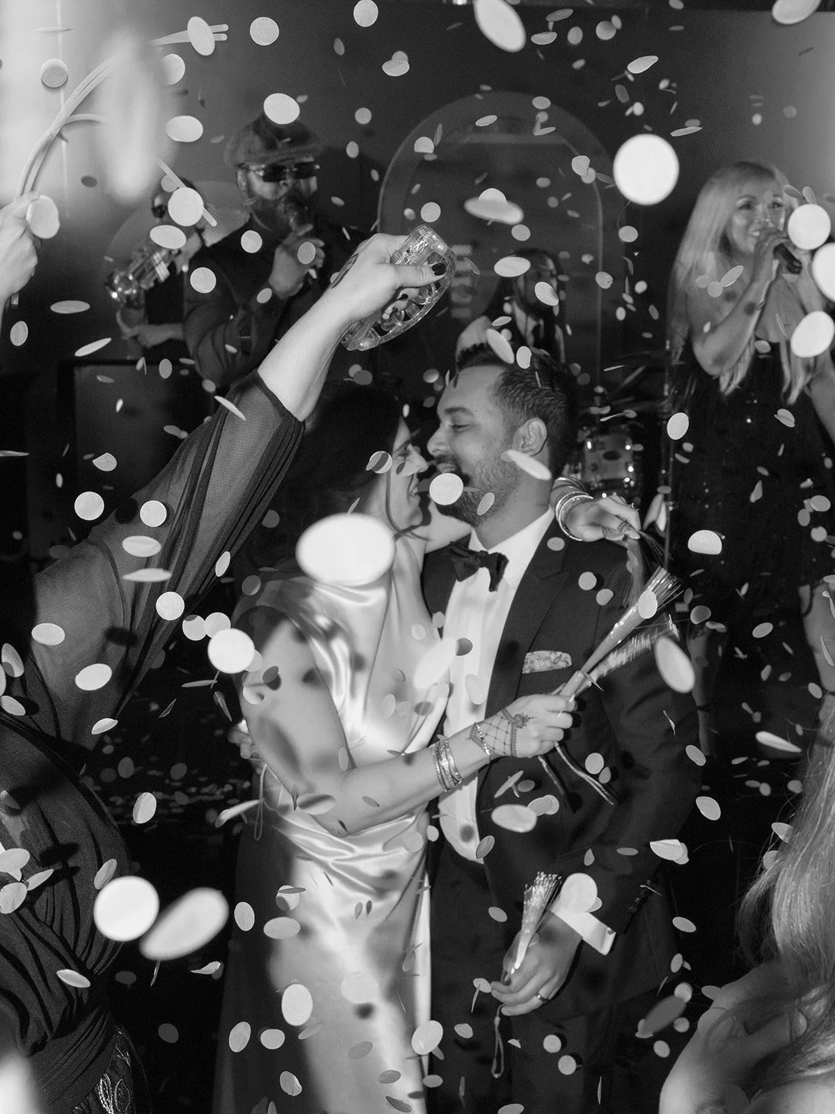 A black and white photo of a newlywed couple celebrating at their wedding reception. They are surrounded by confetti and guests, with one guest holding a party popper. The bride and groom are close, smiling and about to kiss, with the bride holding a bouquet. The background features other guests, including a woman singing.
