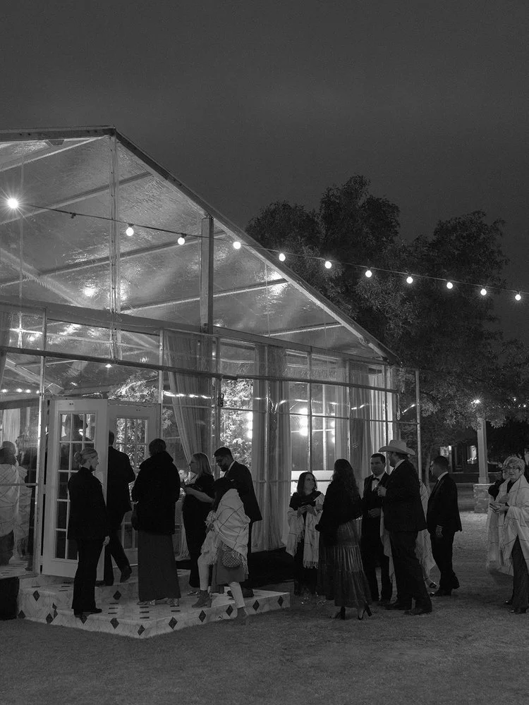 People dressed in formal attire gathering outside a glass greenhouse decorated with string lights at night.
