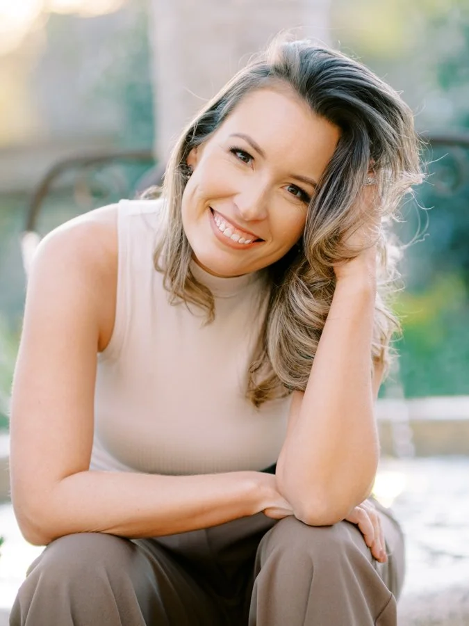 A woman with wavy, shoulder-length hair smiling and looking at the camera, sitting outdoors on a sunny day.