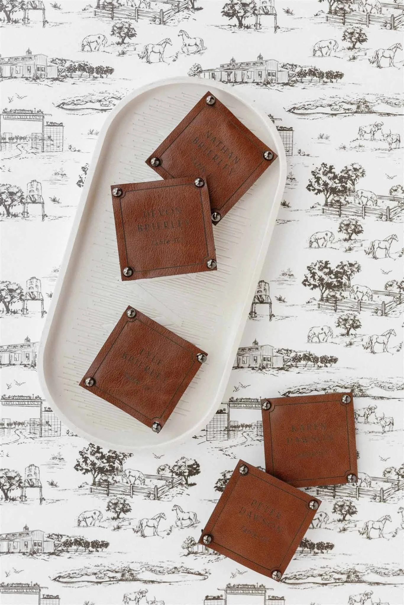 White tray with four brown leather coasters on a black and white countryside-themed wallpaper background featuring horses, trees, farm buildings, and fences.