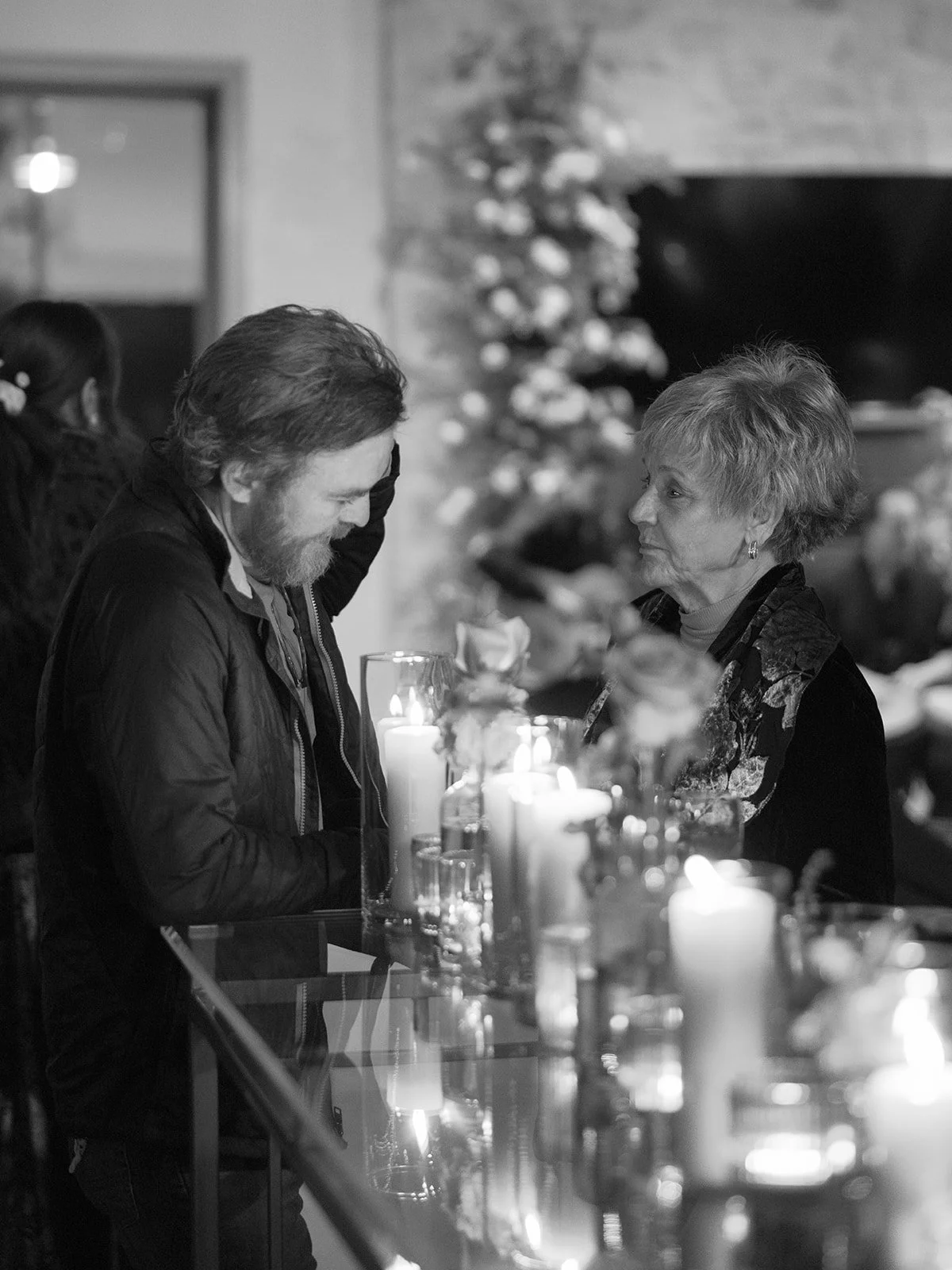 A man with long hair and a beard leans on a bar counter, smiling, as he faces an older woman with short hair. The scene is decorated for Christmas with a tree in the background and lit candles on the counter.