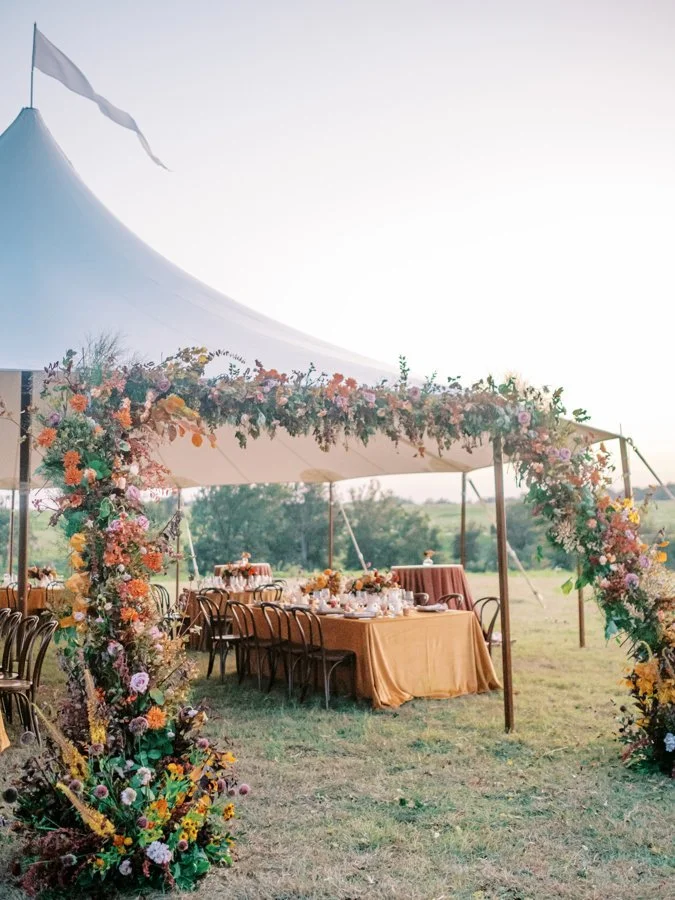 Outdoor wedding reception setup with a large tent, decorated with floral arch and tables with mustard-colored tablecloths, in a grassy field during sunset.