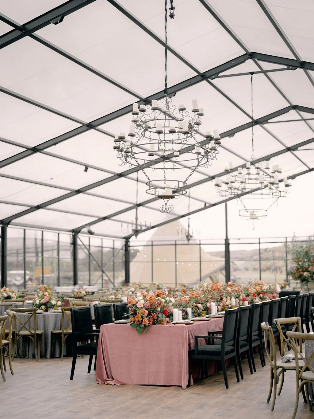 Elegant indoor wedding reception hall with long pink-draped table, floral centerpieces, black chairs, and chandeliers hanging from the ceiling.