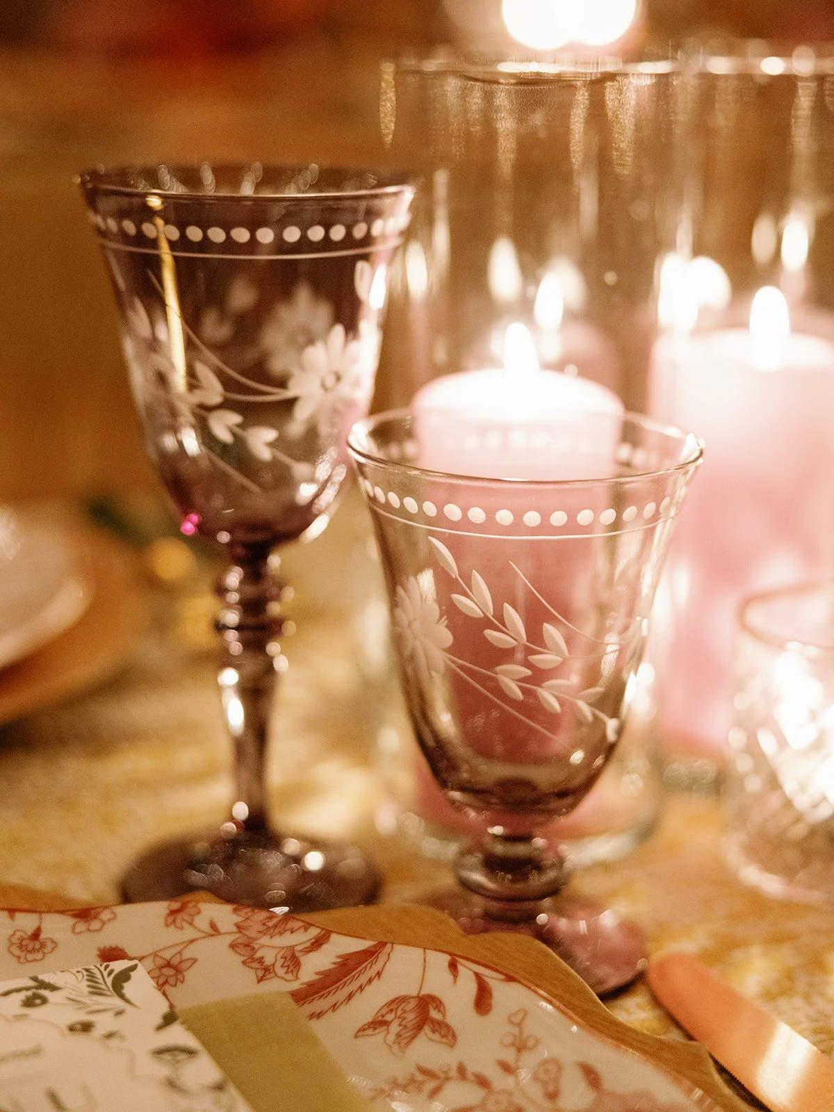 Close-up of two lit pink candles in glass holders with white floral patterns, set on a table with other candles and glassware.