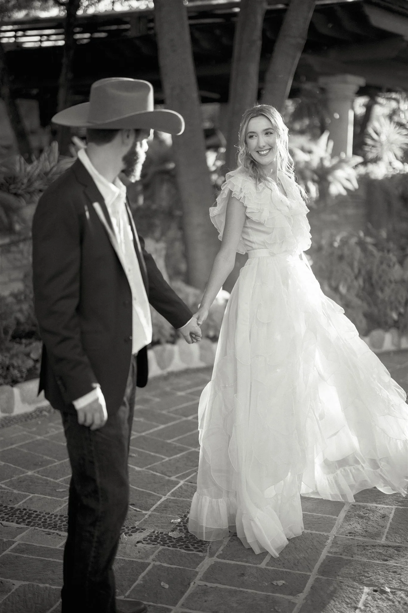 A couple holding hands, walking outdoors, with the woman smiling brightly. The woman is wearing a flowing white dress with ruffles, and the man is dressed in a dark suit with a cowboy hat.