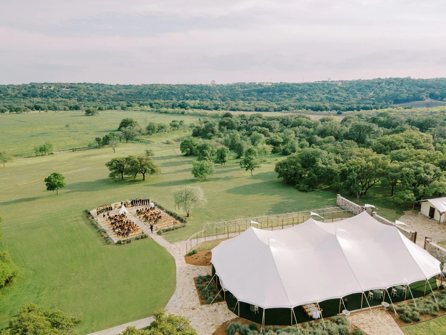 A wedding ceremony outdoors on a grassy field with a white tent nearby, guests seated facing a small altar, surrounded by lush trees and open green landscape