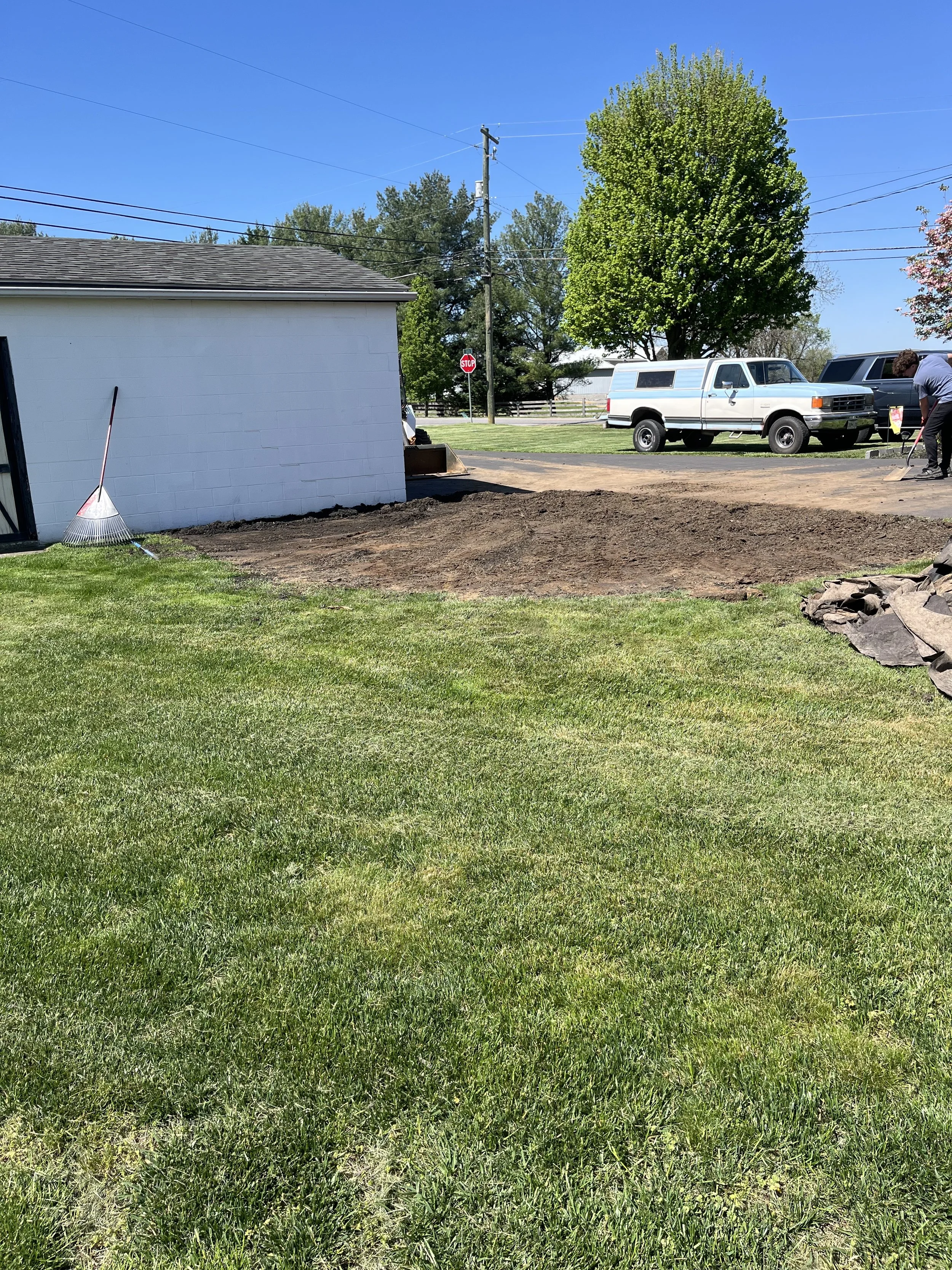 A before image of backyard with a newly tilled dirt area, a white building on the left, a large leafy green tree in the background, a white work truck, and a person raking in the distance on a sunny day.