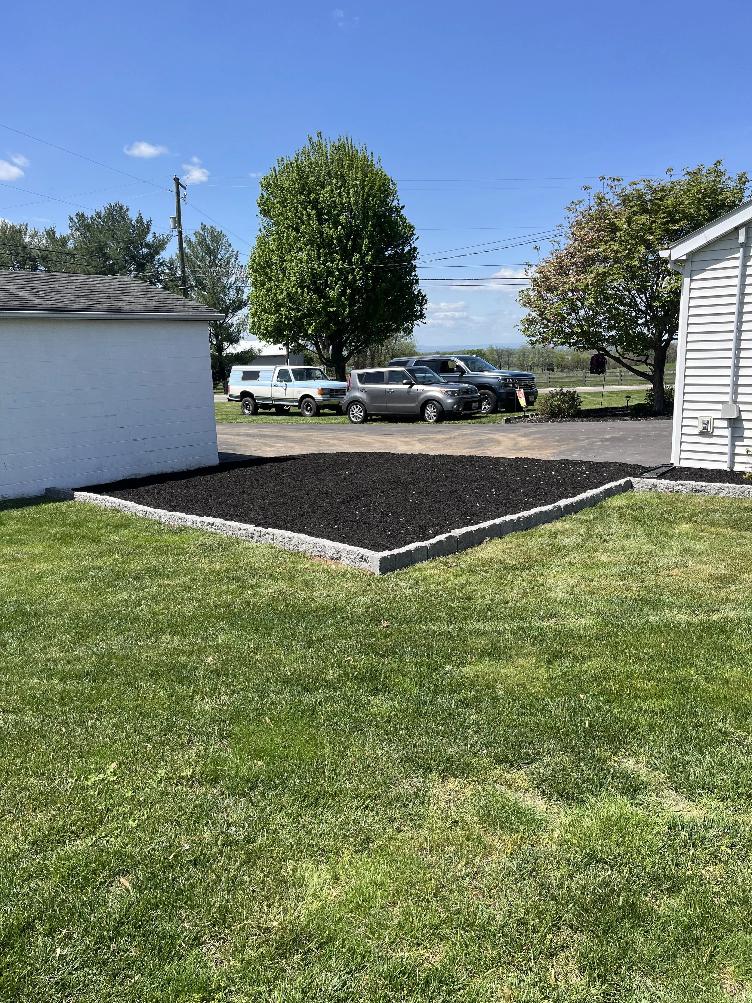 An after image of freshly landscaped garden bed with dark mulch, bordered by gray stones, next to a white house with a white garage and a car parked on the driveway, with two trees and a clear blue sky in the background.