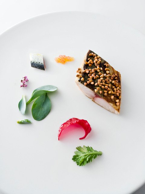 A minimalist gourmet dish displayed on a white plate, featuring a piece of fish with a crunchy topping, various herbs, and small vegetable garnishes.