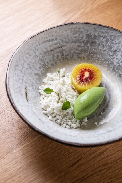 A ceramic bowl containing a scoop of green sorbet, a halved piece of starfruit, and a small garnish of microgreens on a bed of white powder, placed on a wooden surface.