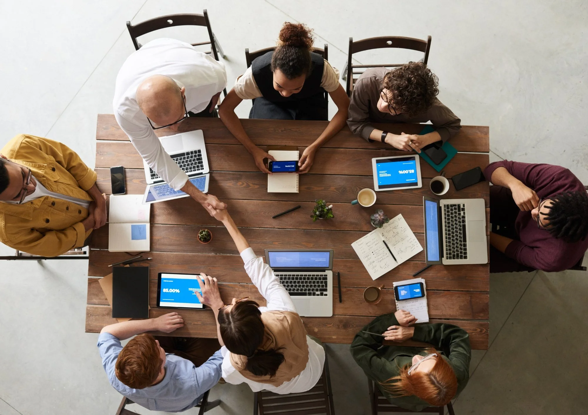 A top-down view of eight people gathered around a wooden conference table, shaking hands and working on laptops, tablets, and smartphones. The table has notebooks, plants, and coffee cups.