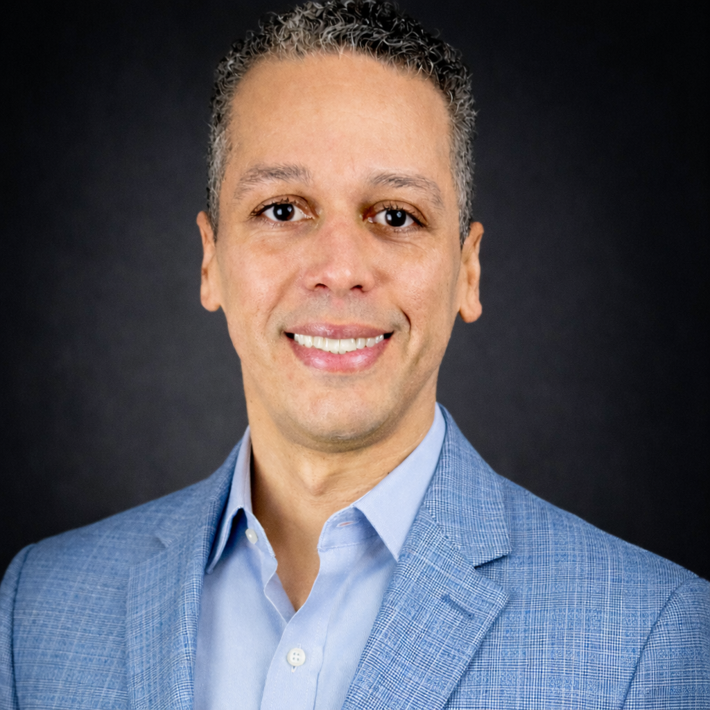 A portrait of Pablo Fana, executive advisor smiling in a light blue suit jacket and shirt against a black background.