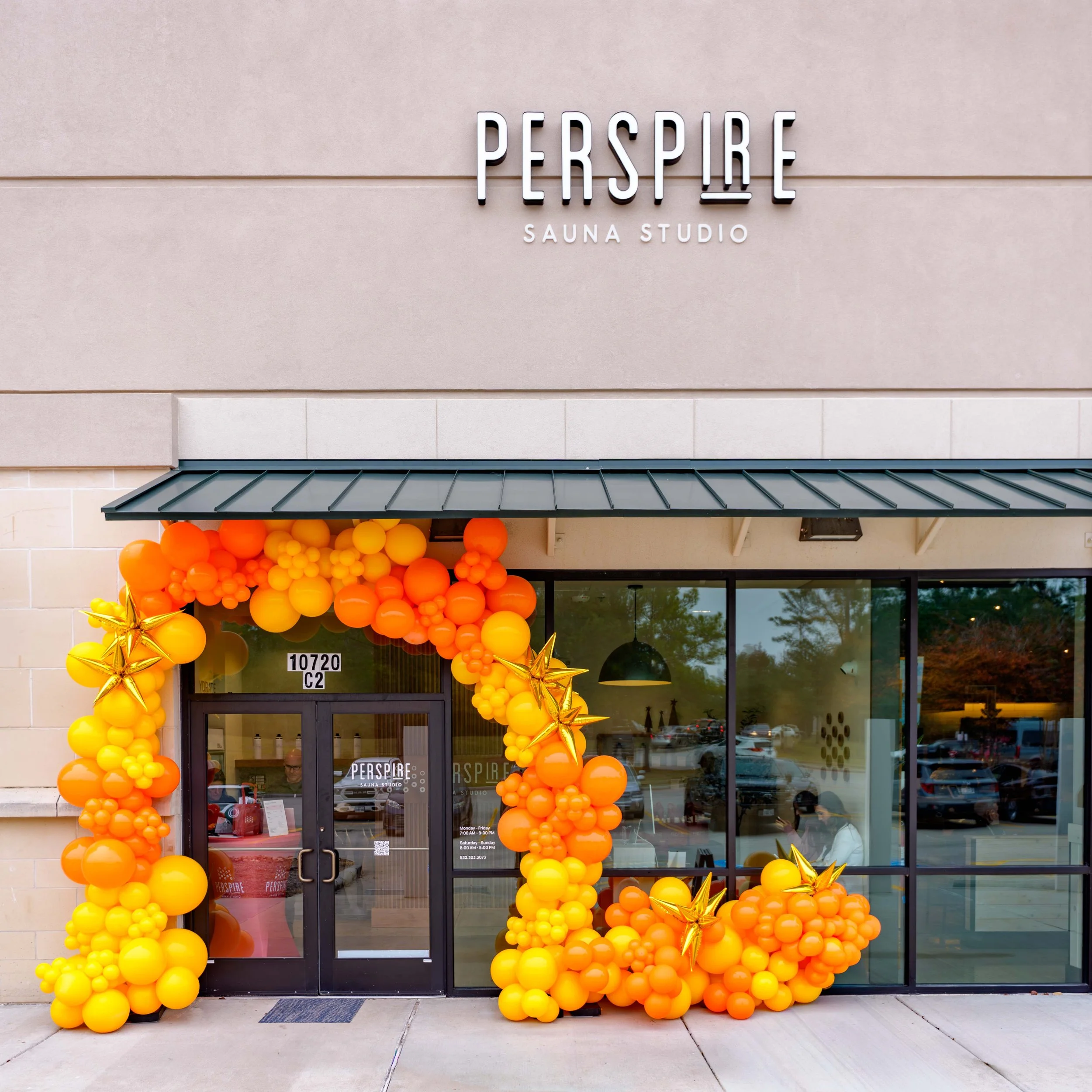 Commercial photography of a storefront featuring orange and yellow balloon decorations around the entrance, with “PERSPIRE Sauna Studio” signage above the door and large display windows.
