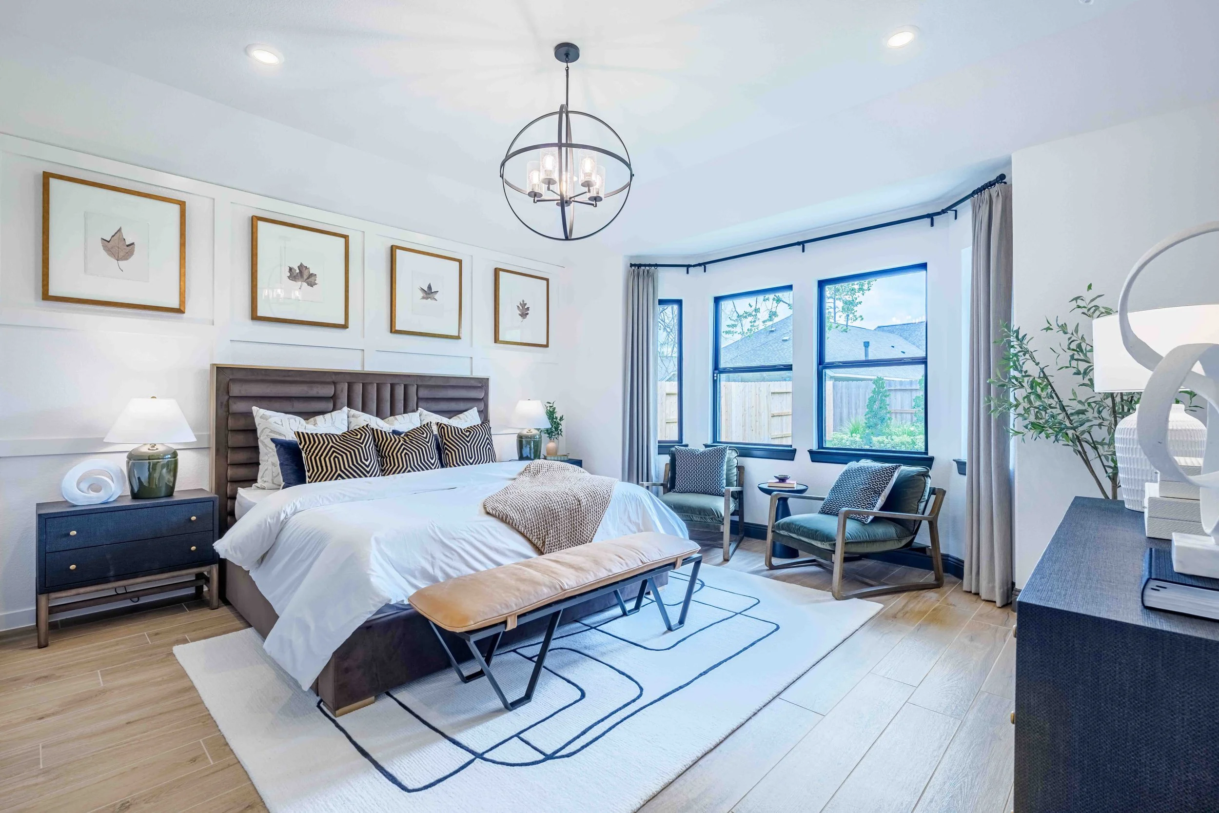 A bright bedroom photographed for a real estate listing, featuring a king-sized bed, framed botanical artwork, matching bedside lamps, two armchairs near the window, a bench at the foot of the bed, and a dresser styled with decorative accents.