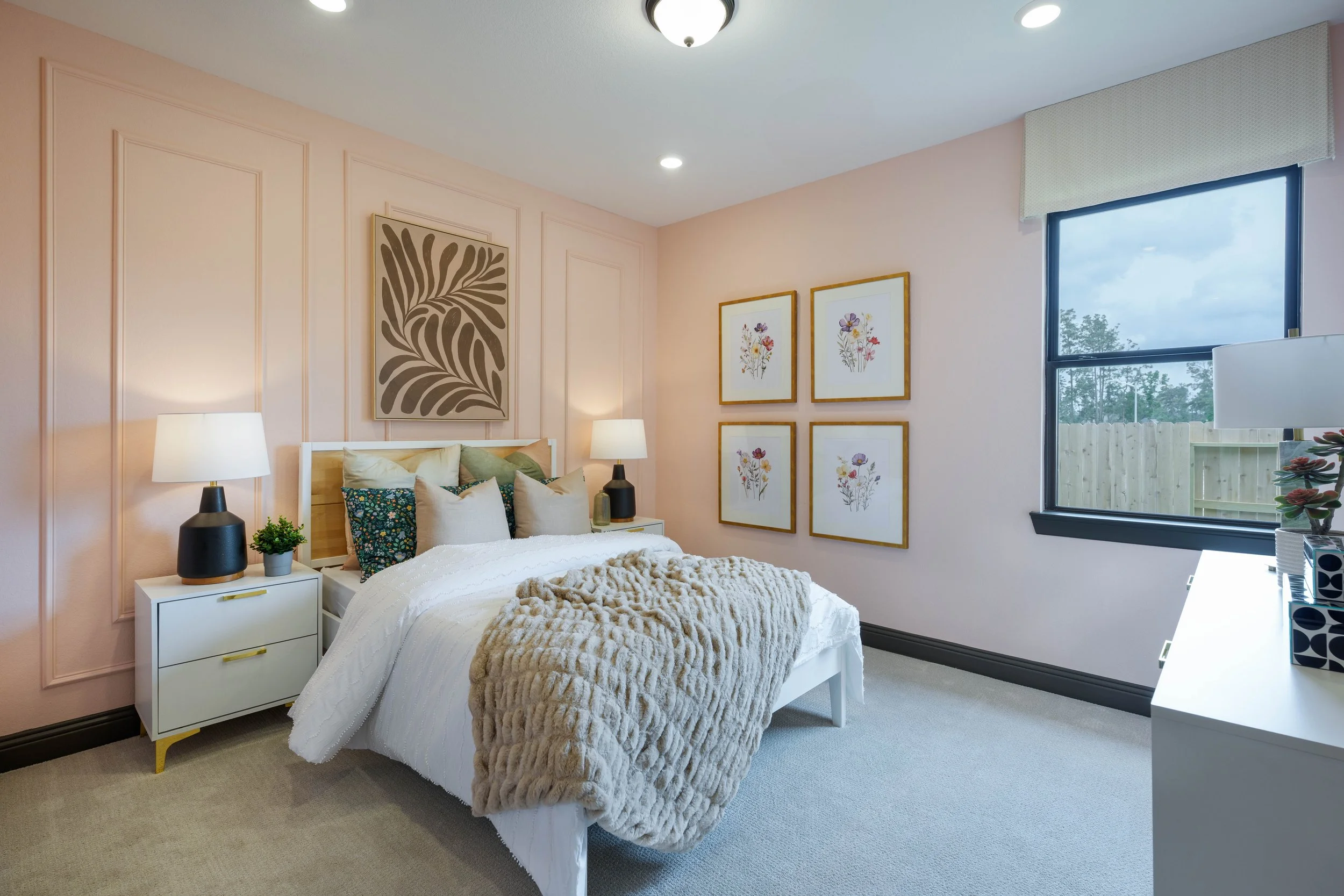A bedroom with light pink walls, a white bed with multiple pillows, a beige chunky blanket, two black and white bedside lamps, framed botanical artwork, a window with a shade, and a white dresser.
