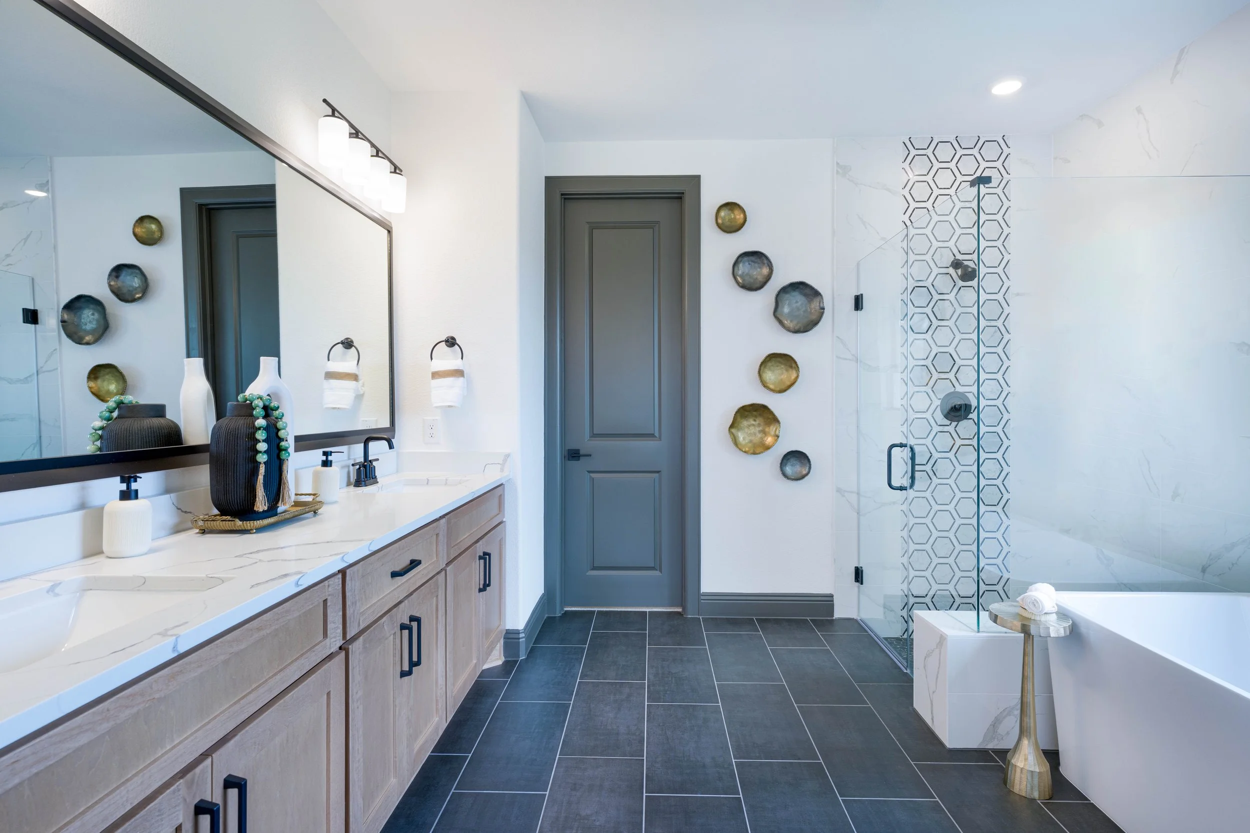 A modern bathroom photographed for a real estate listing, featuring a sleek vanity with a large mirror, decorative vases, abstract wall art, a walk-in shower with patterned tile, a freestanding bathtub, and dark floor tiles.